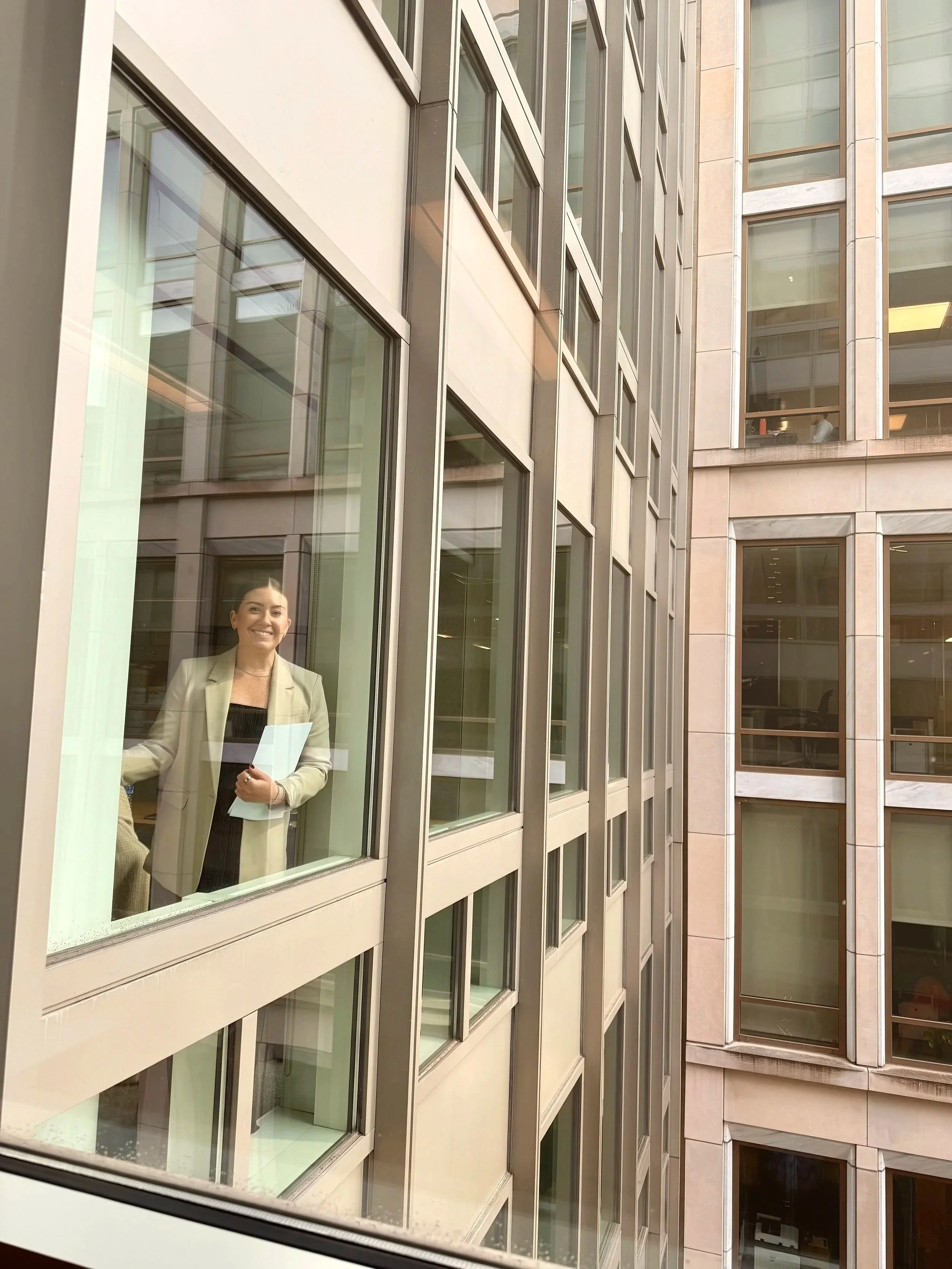 A woman is standing inside an office building, looking out the window and smiling, holding a folder or papers.