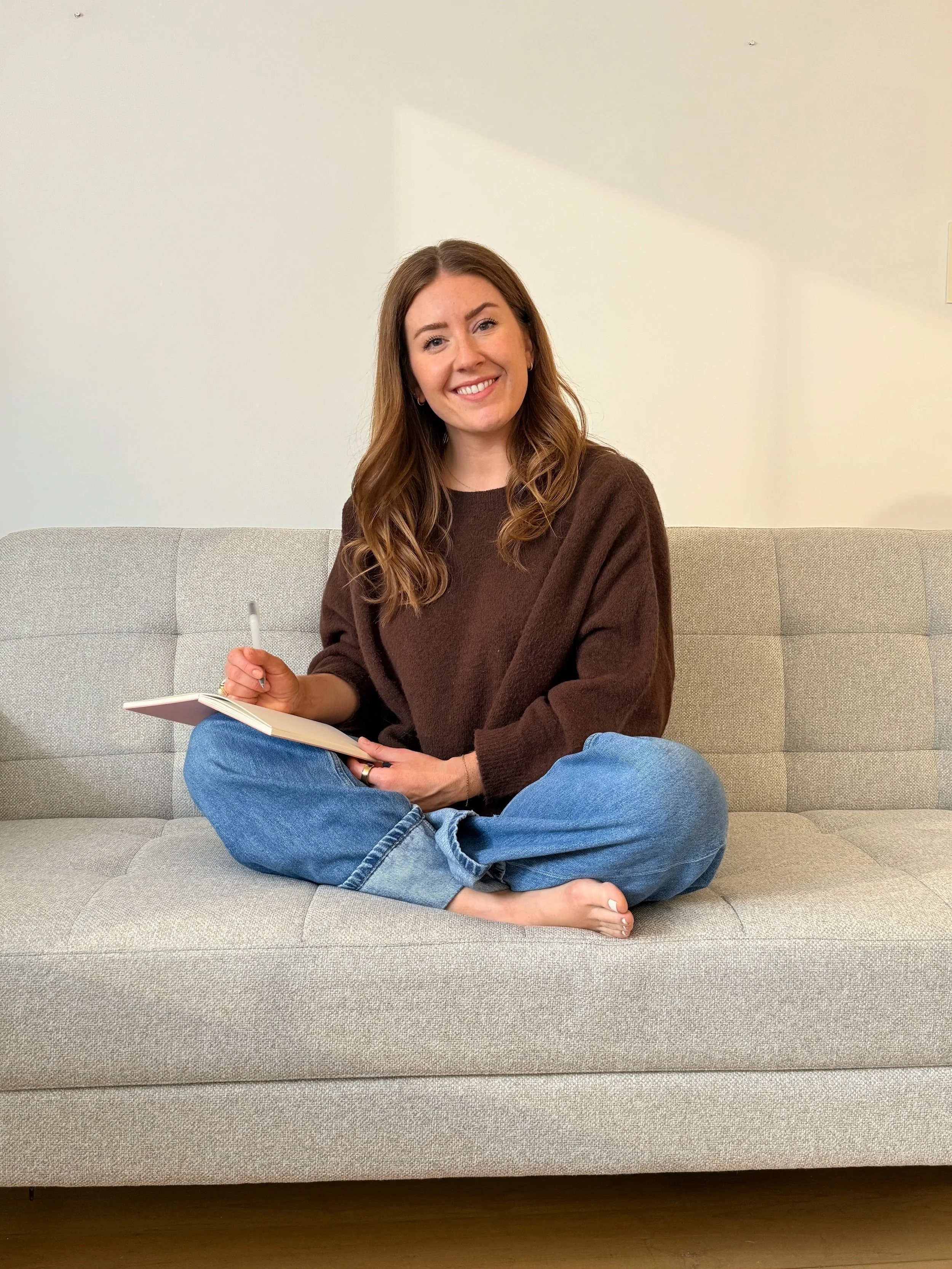 A woman with long brown hair, sitting on a light gray sofa, smiling, holding a pen and an open notebook, dressed in a brown sweater and blue jeans.