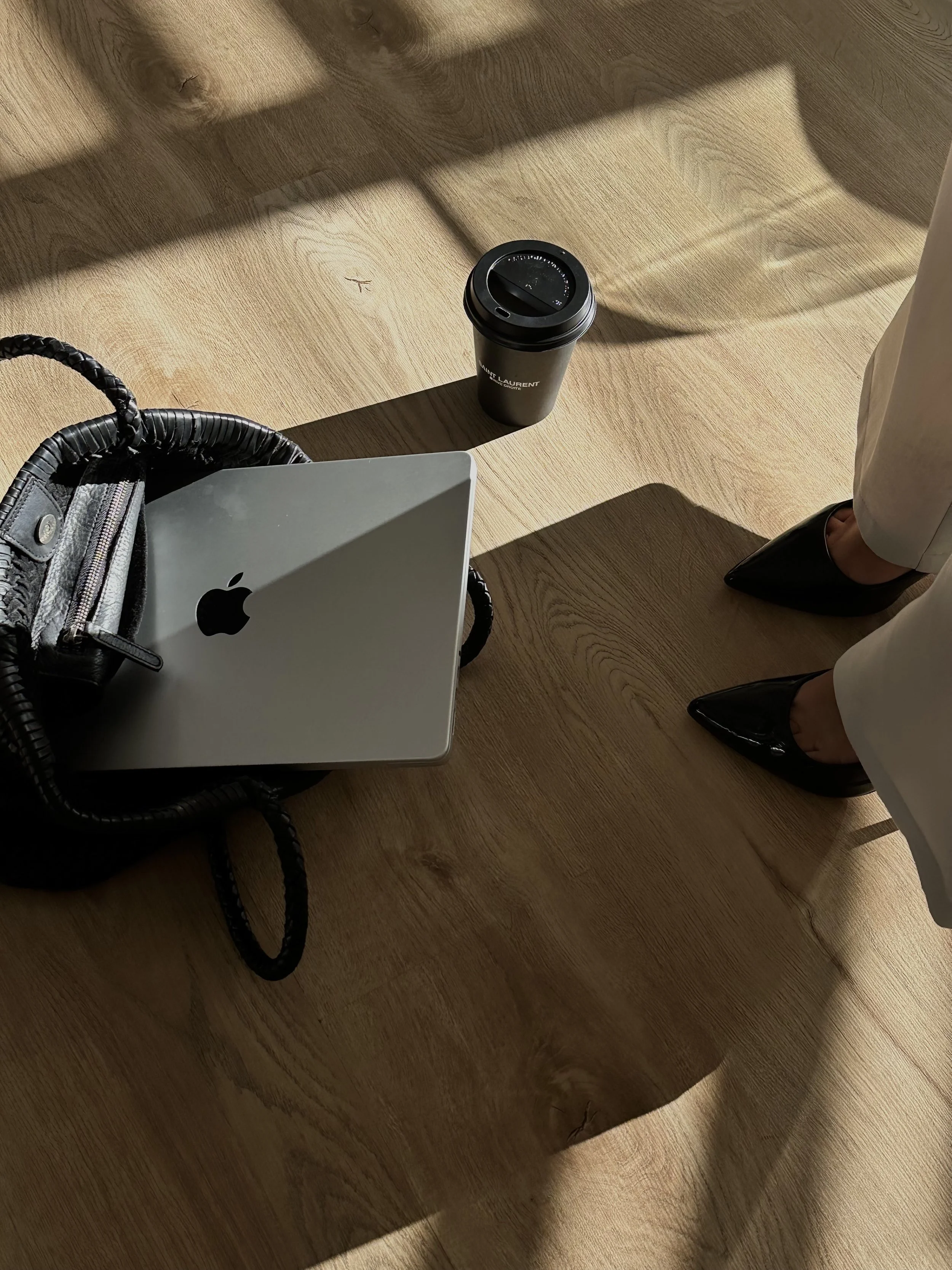 Top-down view of a wooden floor with a black handbag, a closed Apple laptop, a black coffee cup with a lid, and a person's legs wearing white pants and black pointed shoes.