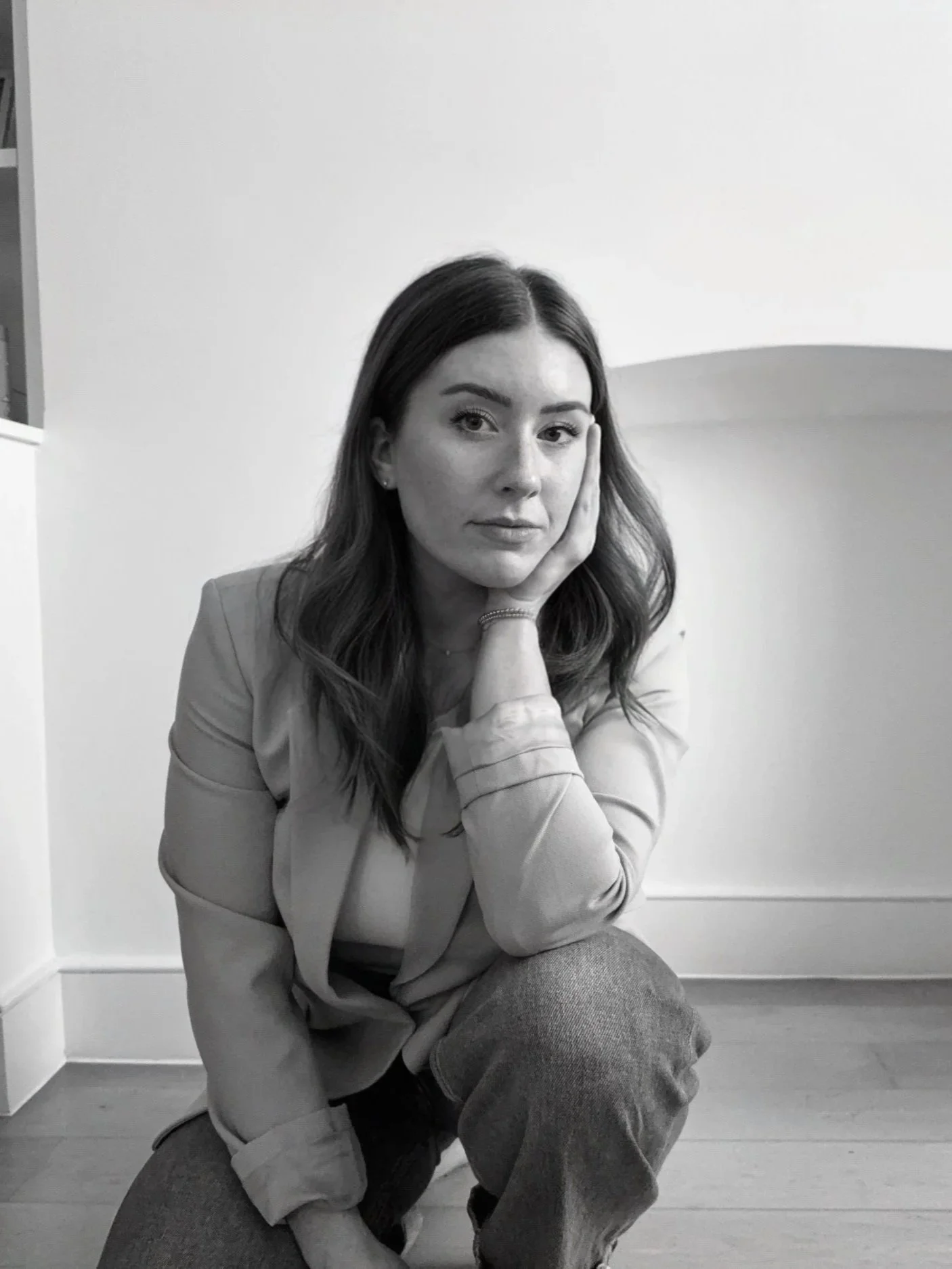 Black-and-white photo of a woman with wavy hair, sitting on the floor, resting her face on her hand, with a neutral expression, indoors against a plain wall.