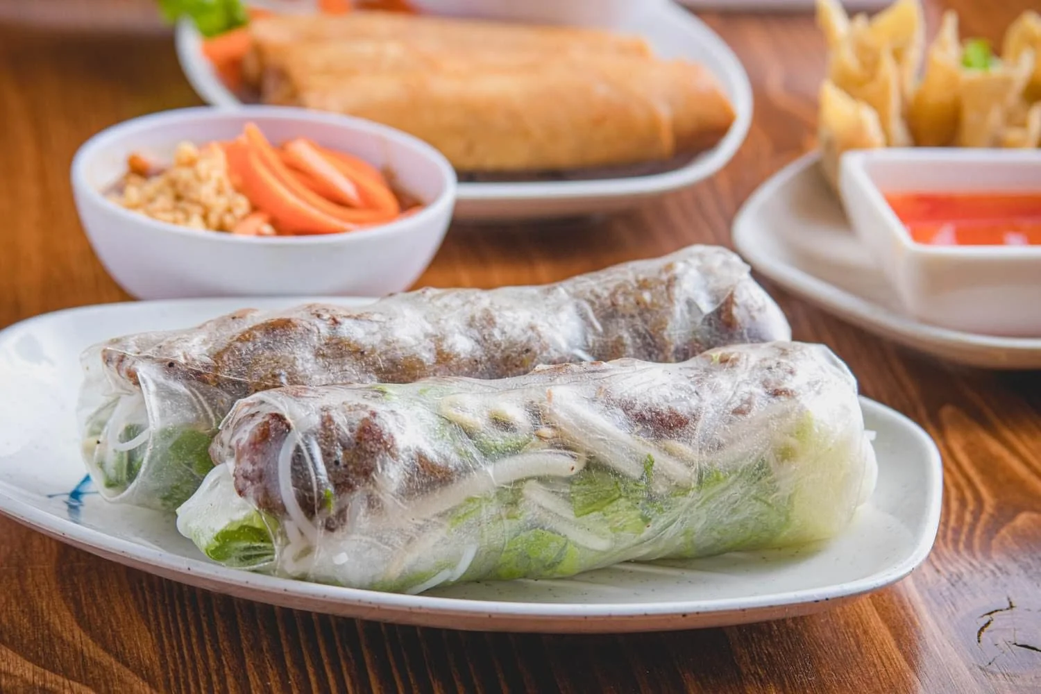 Vietnamese spring rolls on a white plate with lettuce and herbs inside, with side dishes of dipping sauces, chopped peanuts with sliced carrots, fried wontons, and spring rolls on a wooden table.