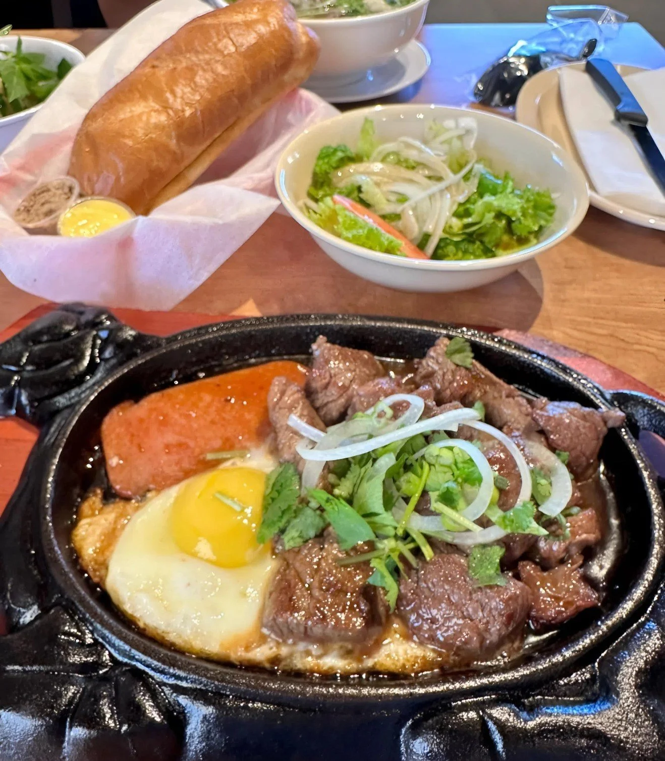 A meal with a sizzling plate of beef topped with onions and cilantro, a fried egg, sliced sausage, a bread roll with butter, a bowl of salad with lettuce and vegetables, and a bowl of soup on a wooden table.