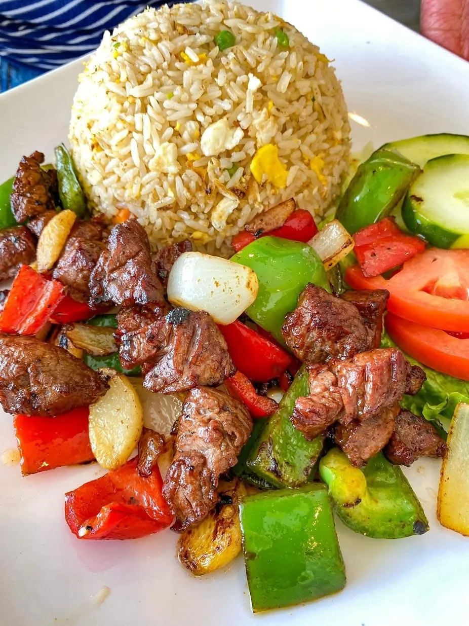 Stir-fried beef with chopped bell peppers, onions, and garlic served with a side of fried rice and sliced cucumbers and tomatoes.