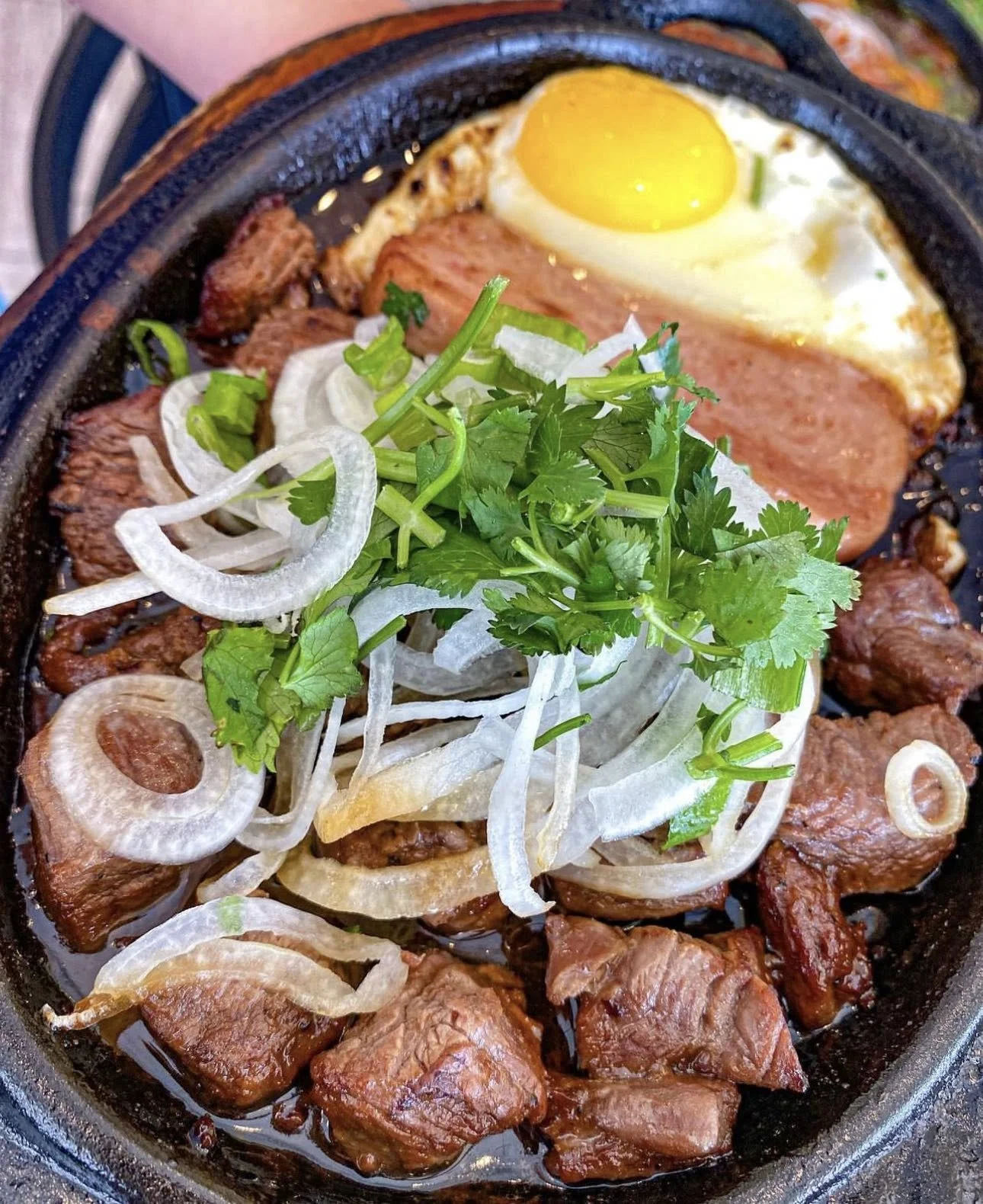 Sizzling steak topped with sliced onions, cilantro, a fried egg, and grilled green chili in a cast iron skillet.