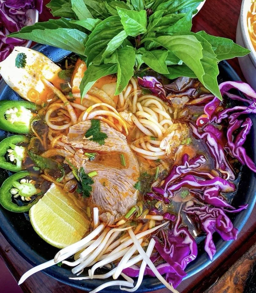 A bowl of Vietnamese pho with slices of beef, noodles, fresh herbs, lime wedge, sliced green chili peppers, and purple cabbage garnished garnished with basil leaves.