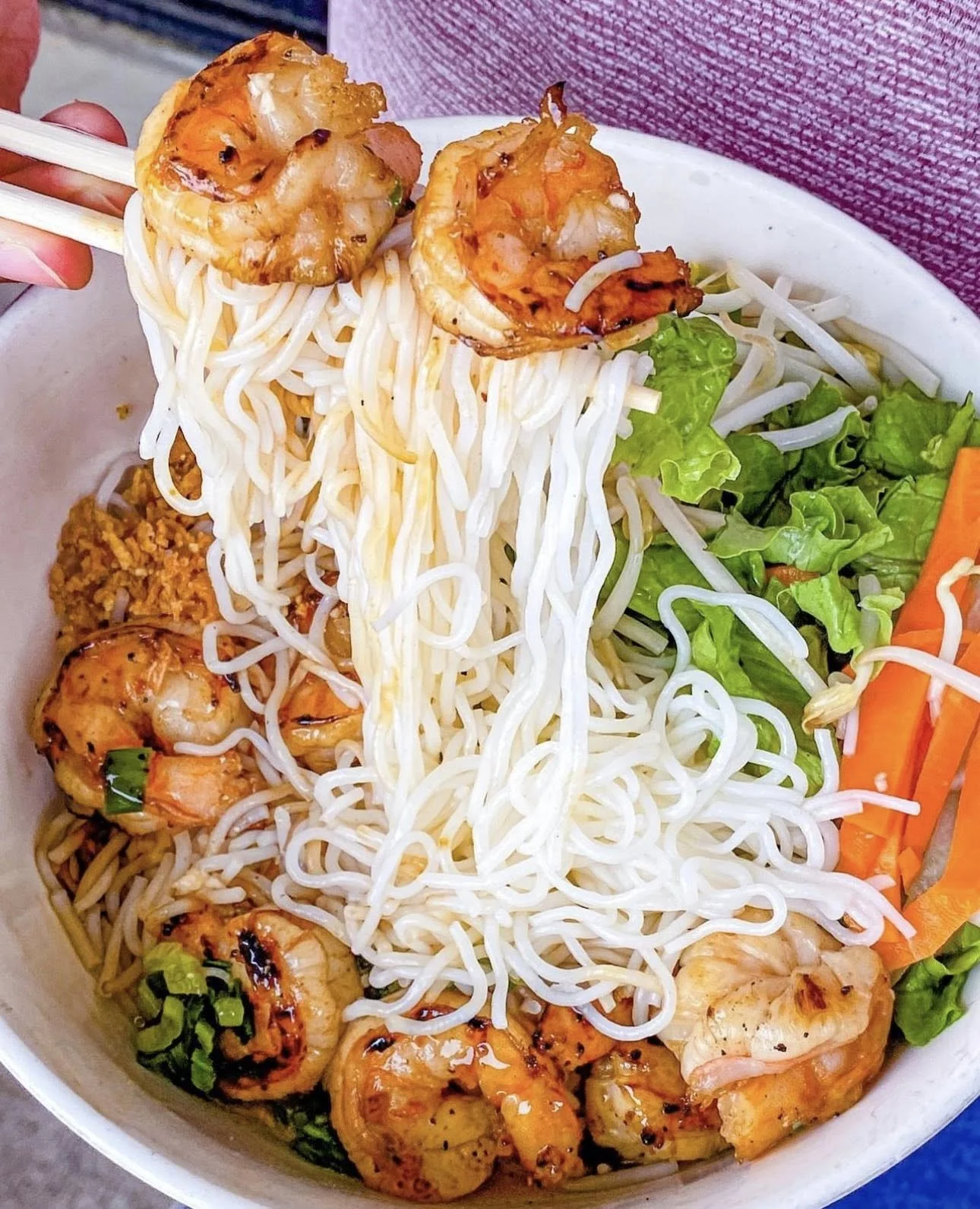 A bowl of Asian stir-fried shrimp with noodles, green lettuce, shredded carrots, and chopped green onions.