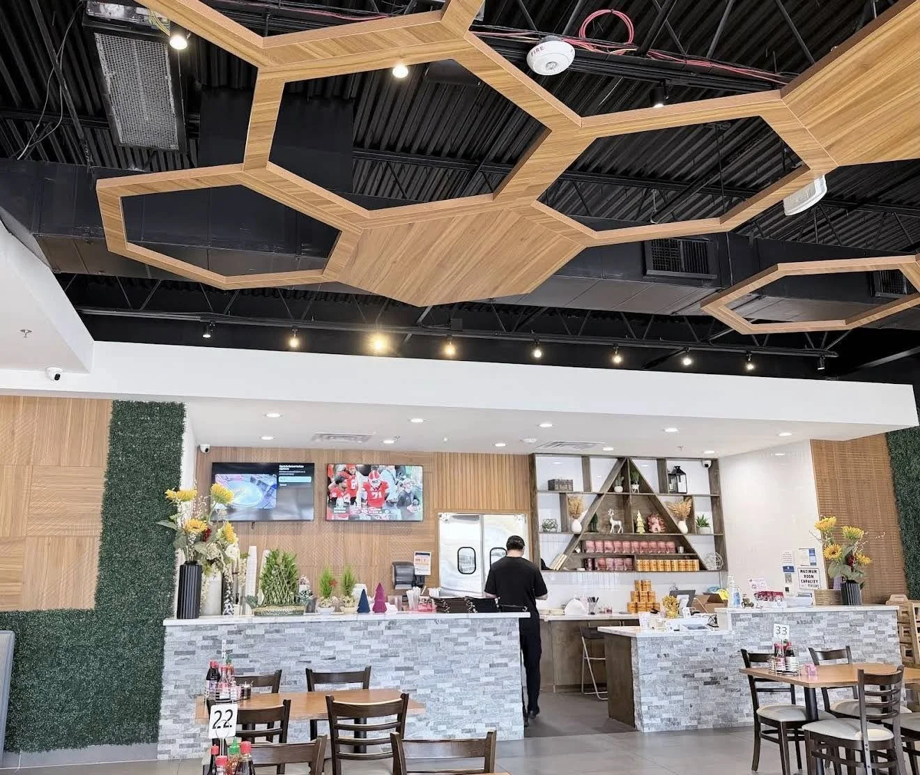 Interior view of a restaurant, featuring a counter with a barista, wooden tables and chairs, decorative shelves, and ceiling with wooden geometric panels and black ceiling, TV screens, and floral arrangements.
