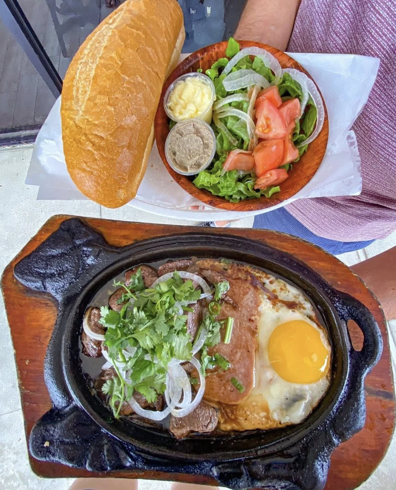 A meal featuring a small cast-iron skillet with cooked beef, a fried egg, and herbs, alongside a wooden bowl of salad with lettuce, tomato, onions, and dressings, and a baguette bread on a white paper-lined tray.