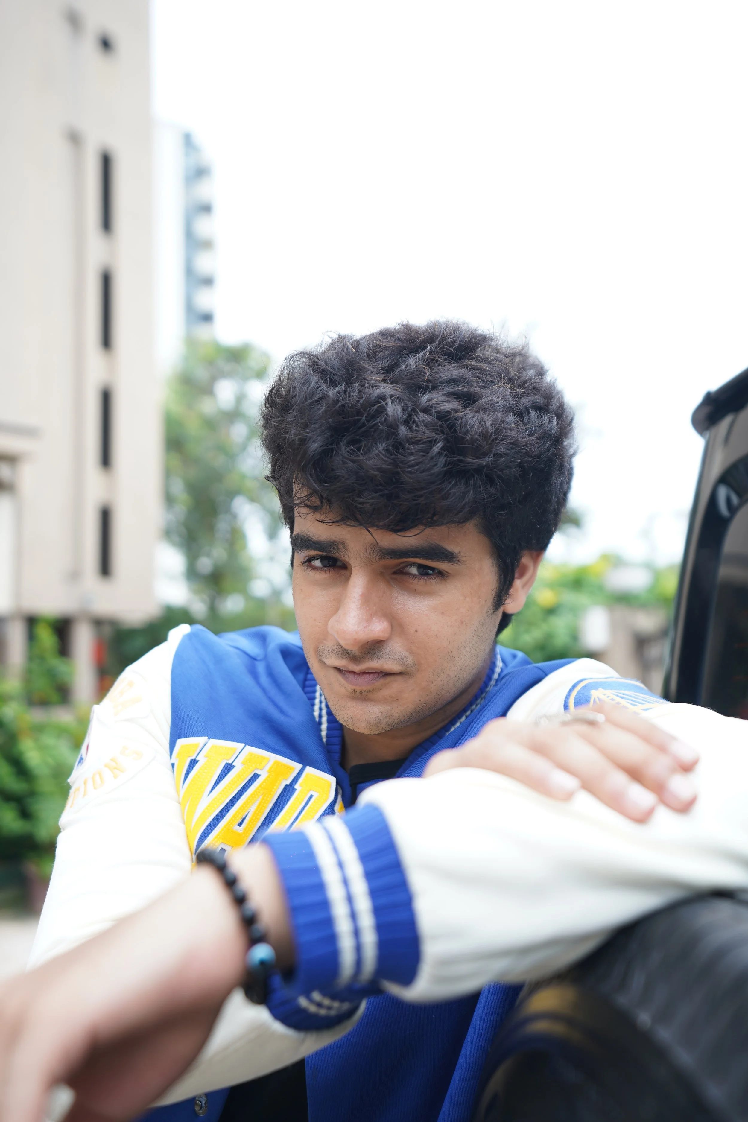 Young man with curly dark hair wearing a blue and white jacket, leaning on a car with a serious expression, outdoors during daytime.