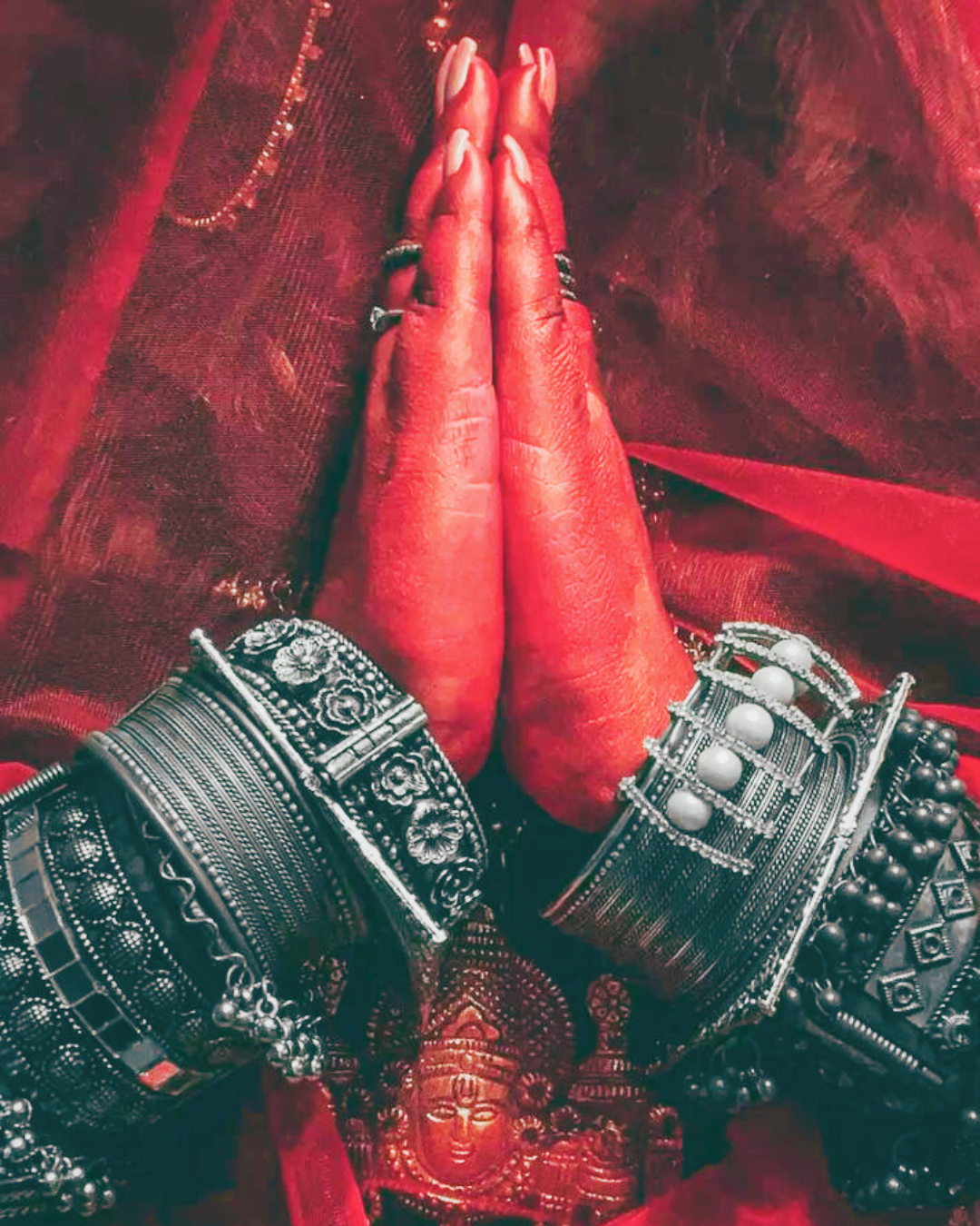 Hands pressed together in a prayer gesture, adorned with jewelry, against a red fabric background.
