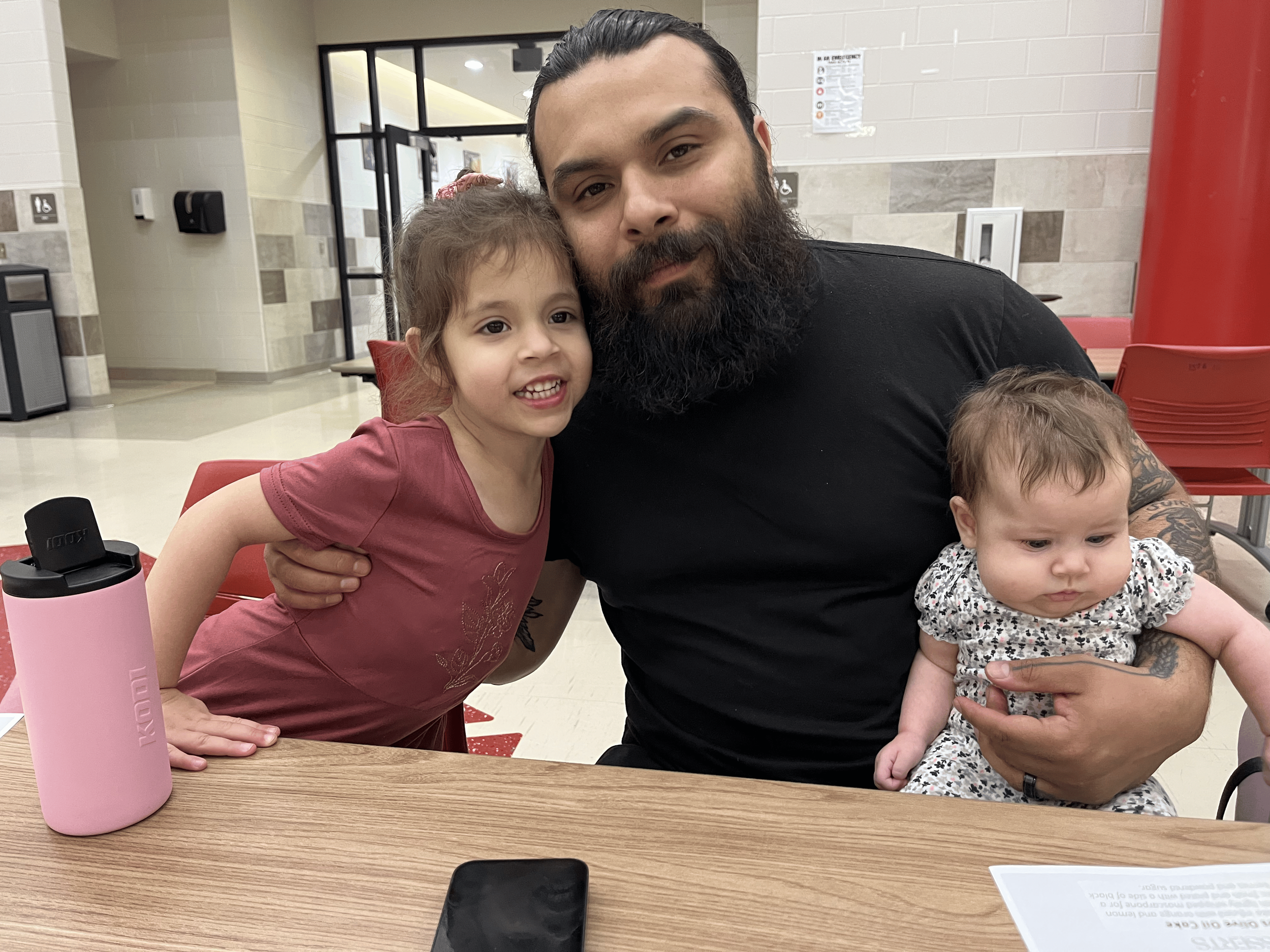 A man with a beard sitting at a table in a public space, holding a young girl on his lap, with another young girl standing beside him. The man is wearing a black shirt, the girl on his lap is wearing a patterned dress, and the standing girl is wearing a reddish dress. They are smiling and facing the camera. The background shows a tiled wall, a glass door, and chairs, suggesting a cafeteria or waiting area.