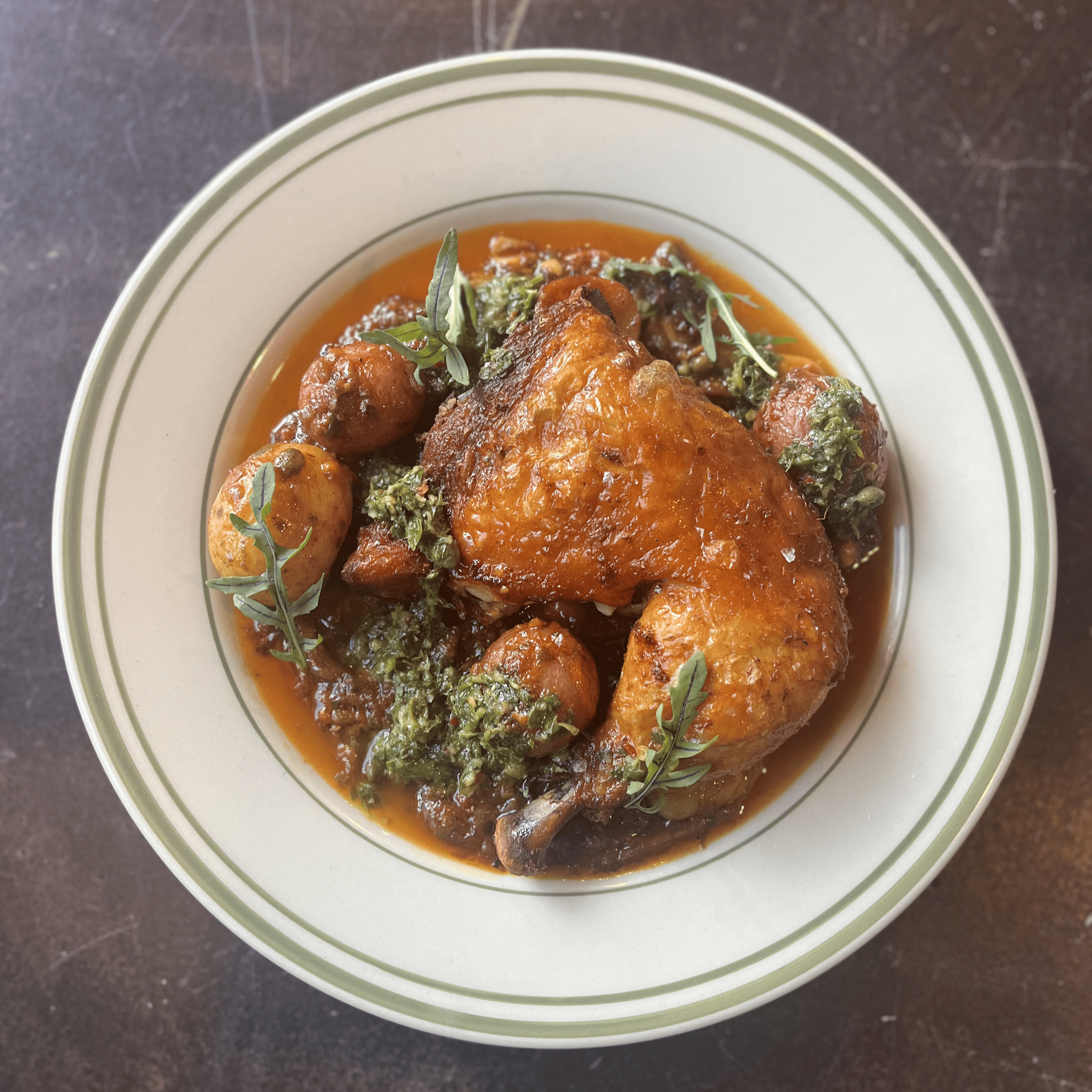 A bowl of cooked chicken drumsticks covered in a brown sauce, garnished with green herbs, and accompanied by roasted potatoes, on a dark surface.