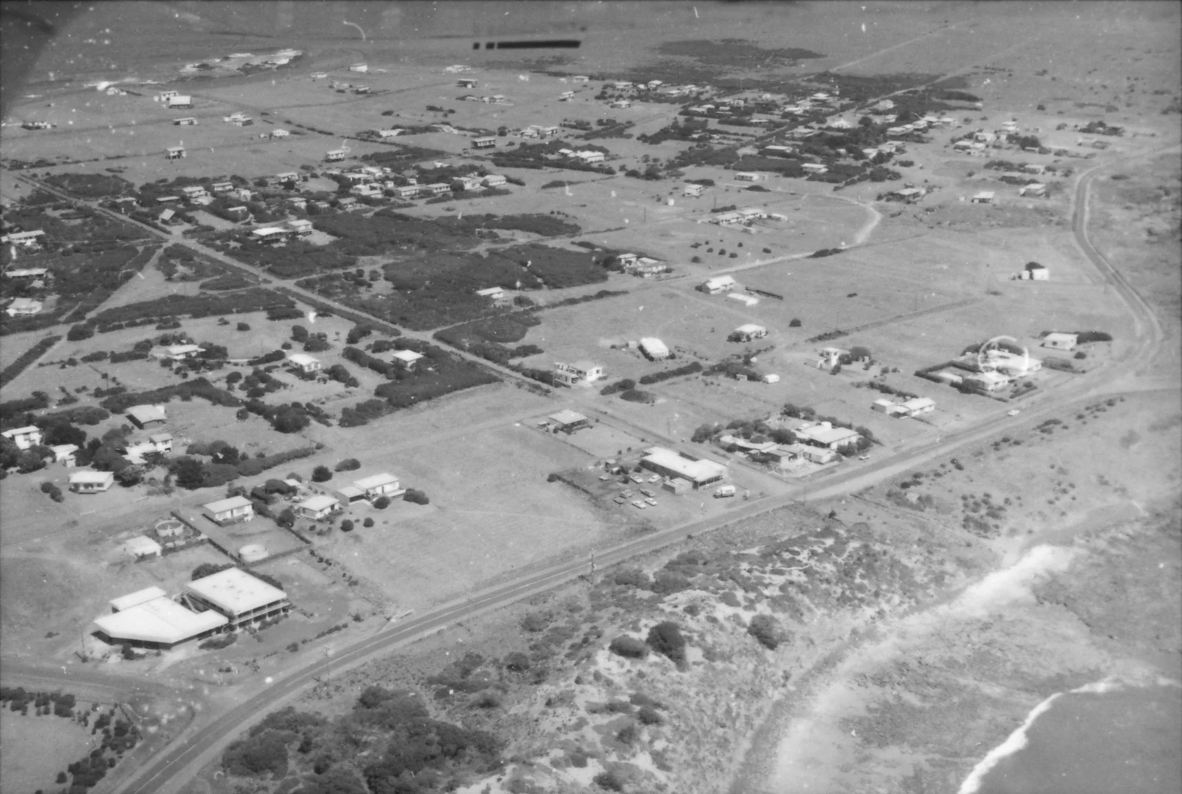Aerial black-and-white photograph of a coastal residential area with houses, roads, and green spaces, near the shoreline with ocean waves.