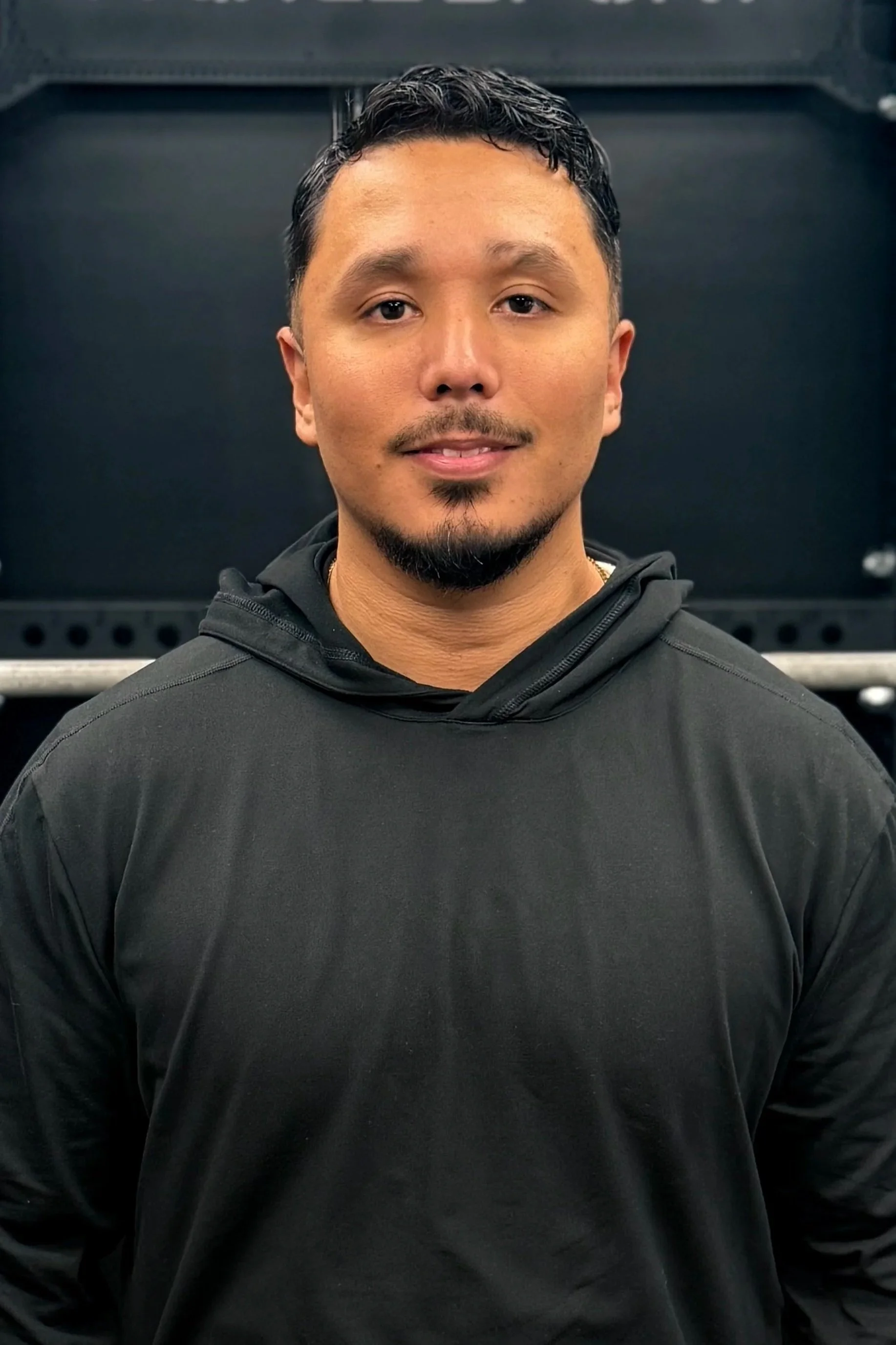A man with short dark hair, facial hair, wearing a black hoodie, standing in front of a dark background with fitness equipment.