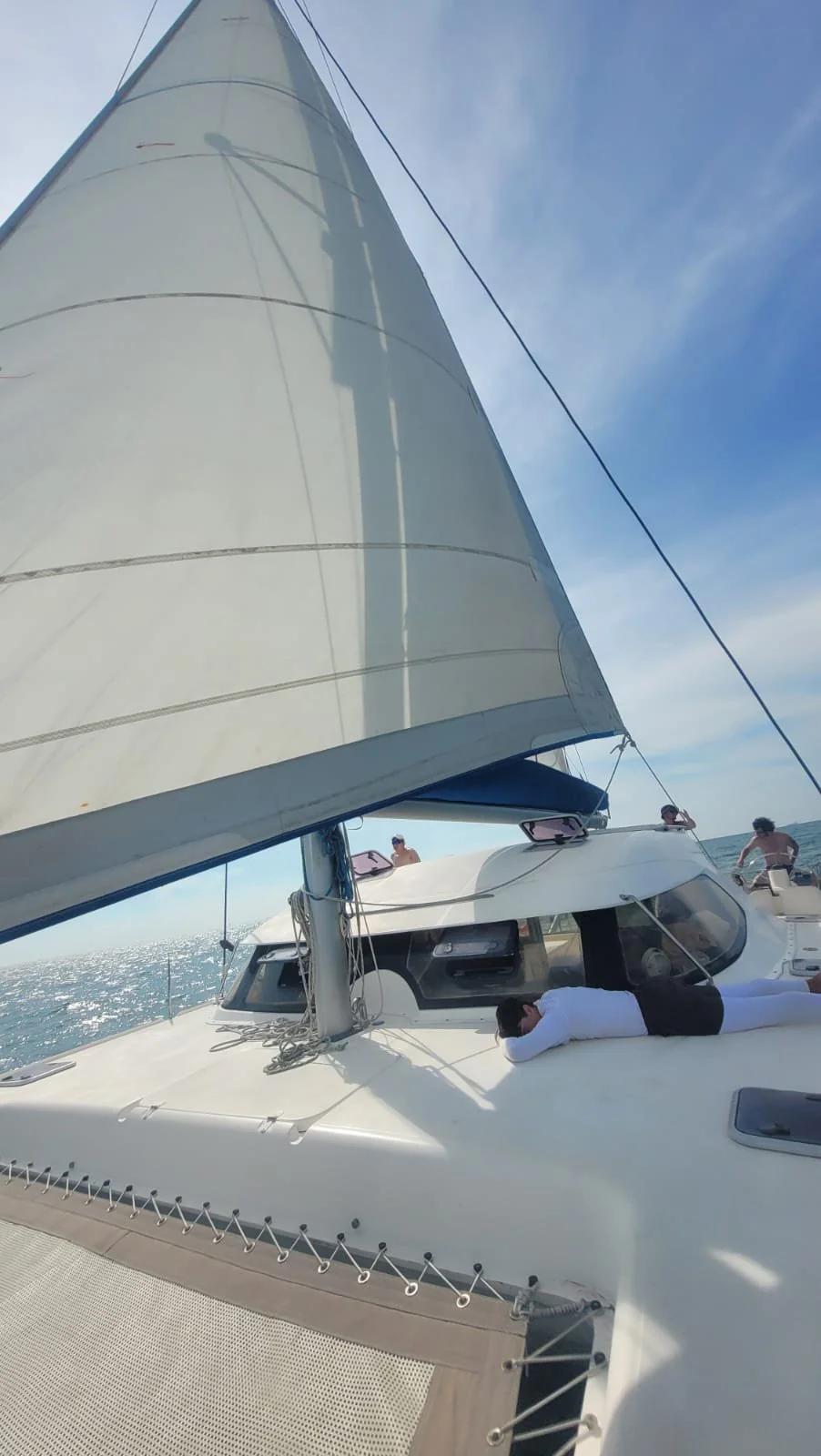 People relaxing on a sailboat during the daytime with the ocean visible in the background. One person is lying on the deck, and others are standing or sitting near the boat's structures.
