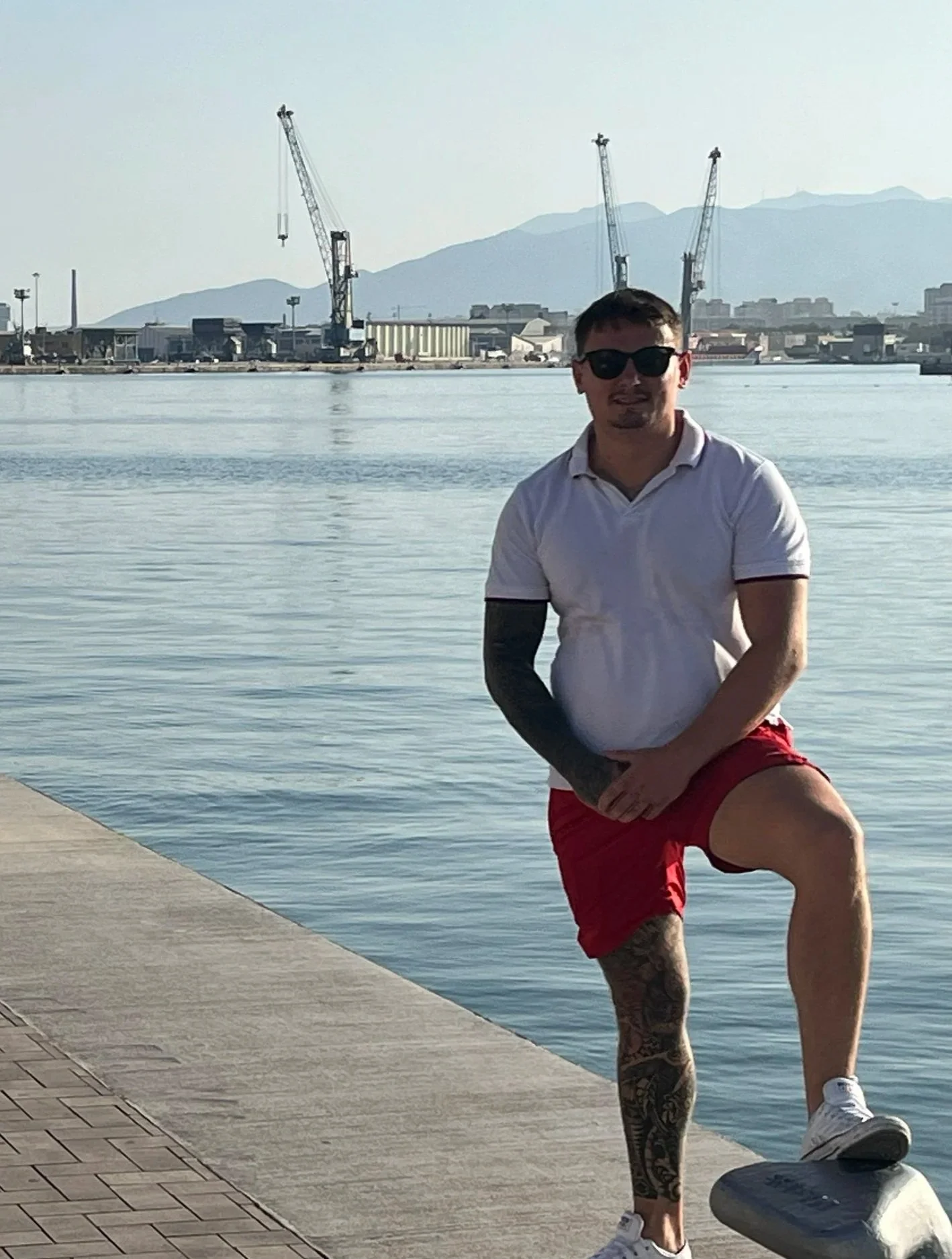 Man with sunglasses, white polo shirt, and red shorts standing on a dock near water, with cranes and mountains in the background.