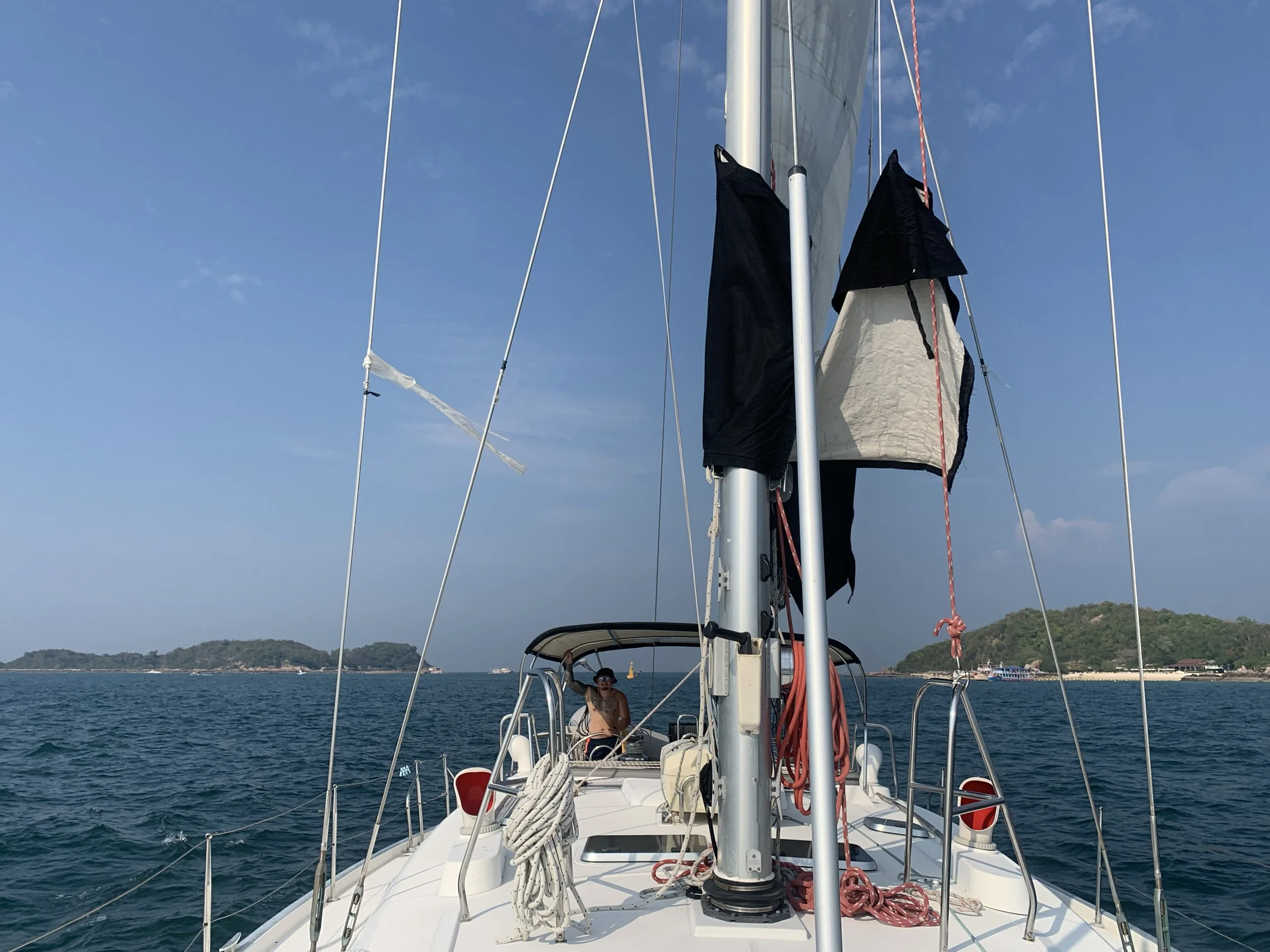 View from the front of a sailboat on the ocean, with a man sitting in the cockpit and islands in the background. The sails are partially down, and ropes and equipment are visible on the boat.