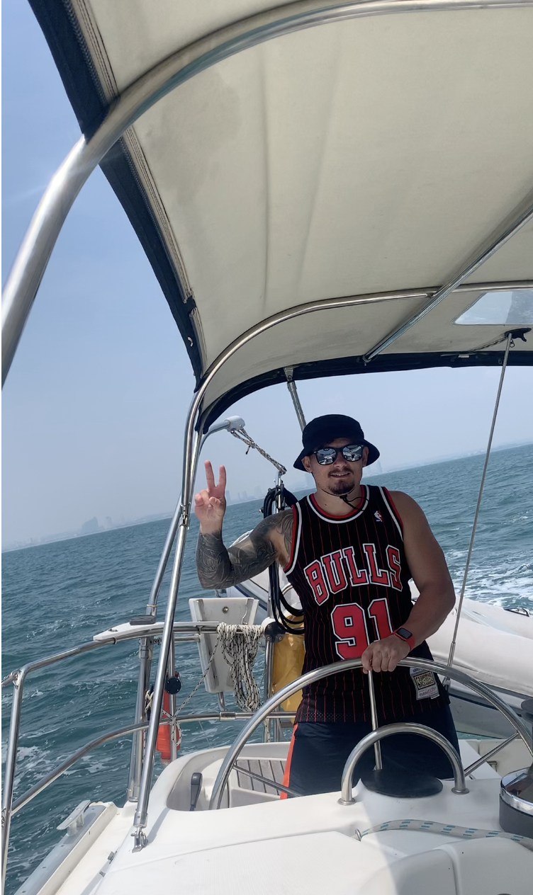 A man wearing a Chicago Bulls jersey, sunglasses, and a bucket hat, standing at the helm of a boat, making a peace sign with his right hand. The boat is on the water under a canopy, with a clear sky and distant city skyline in the background.