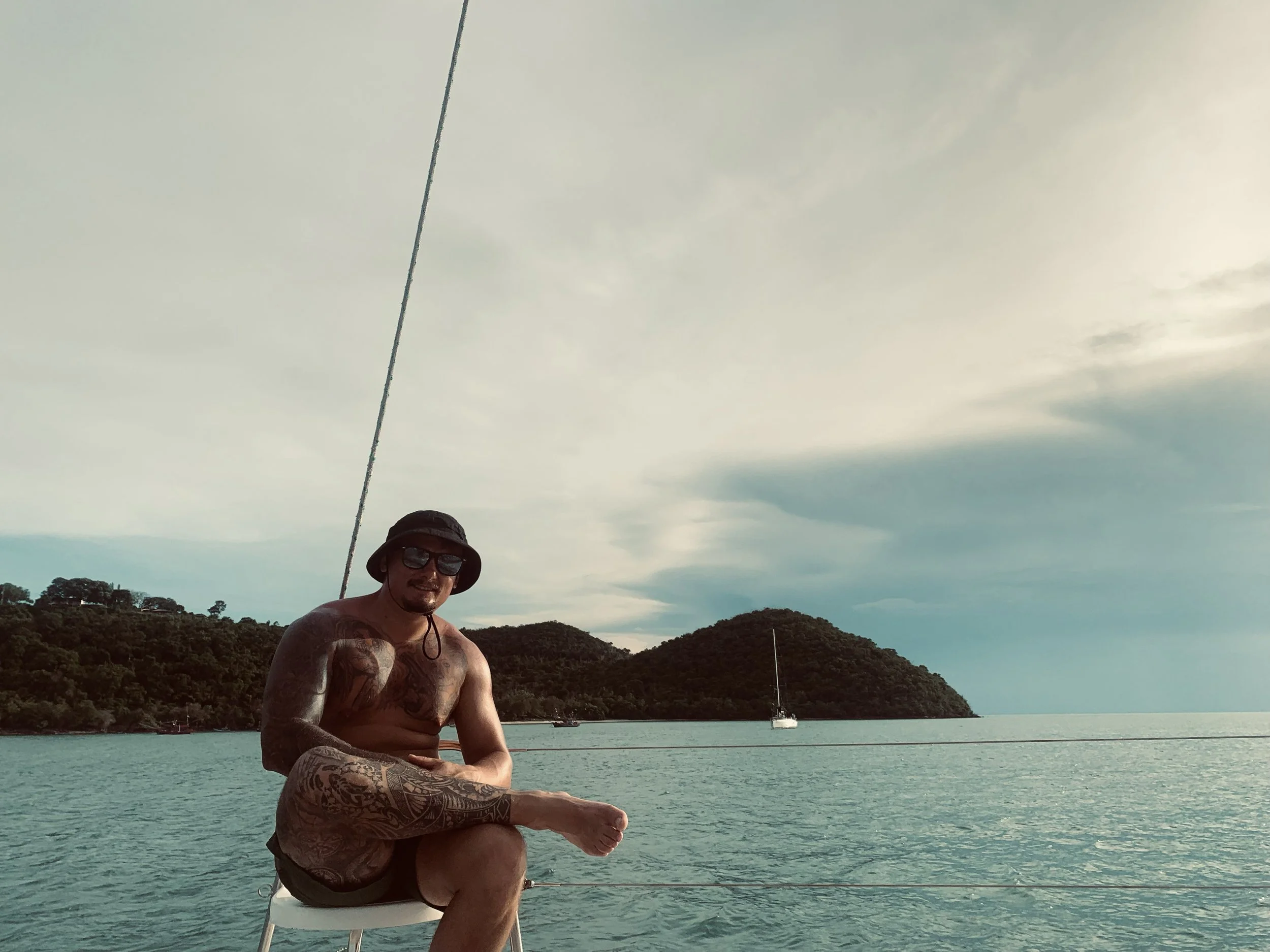 A shirtless man wearing sunglasses, a hat, and shorts sitting on a boat in a body of water, with an island and a sailboat in the background.