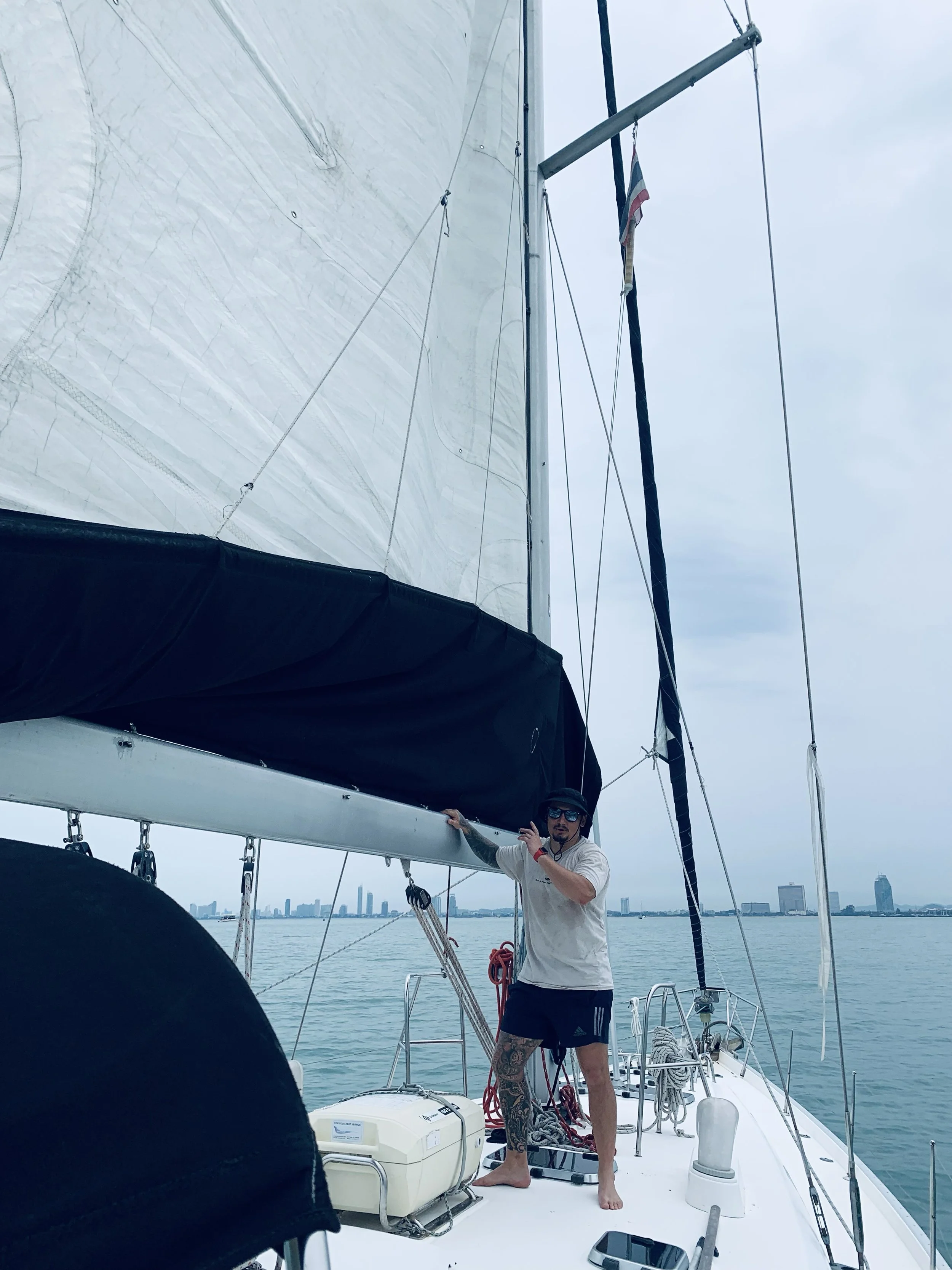 A man on a sailboat adjusting the rigging, with water and city skyline in the distance.