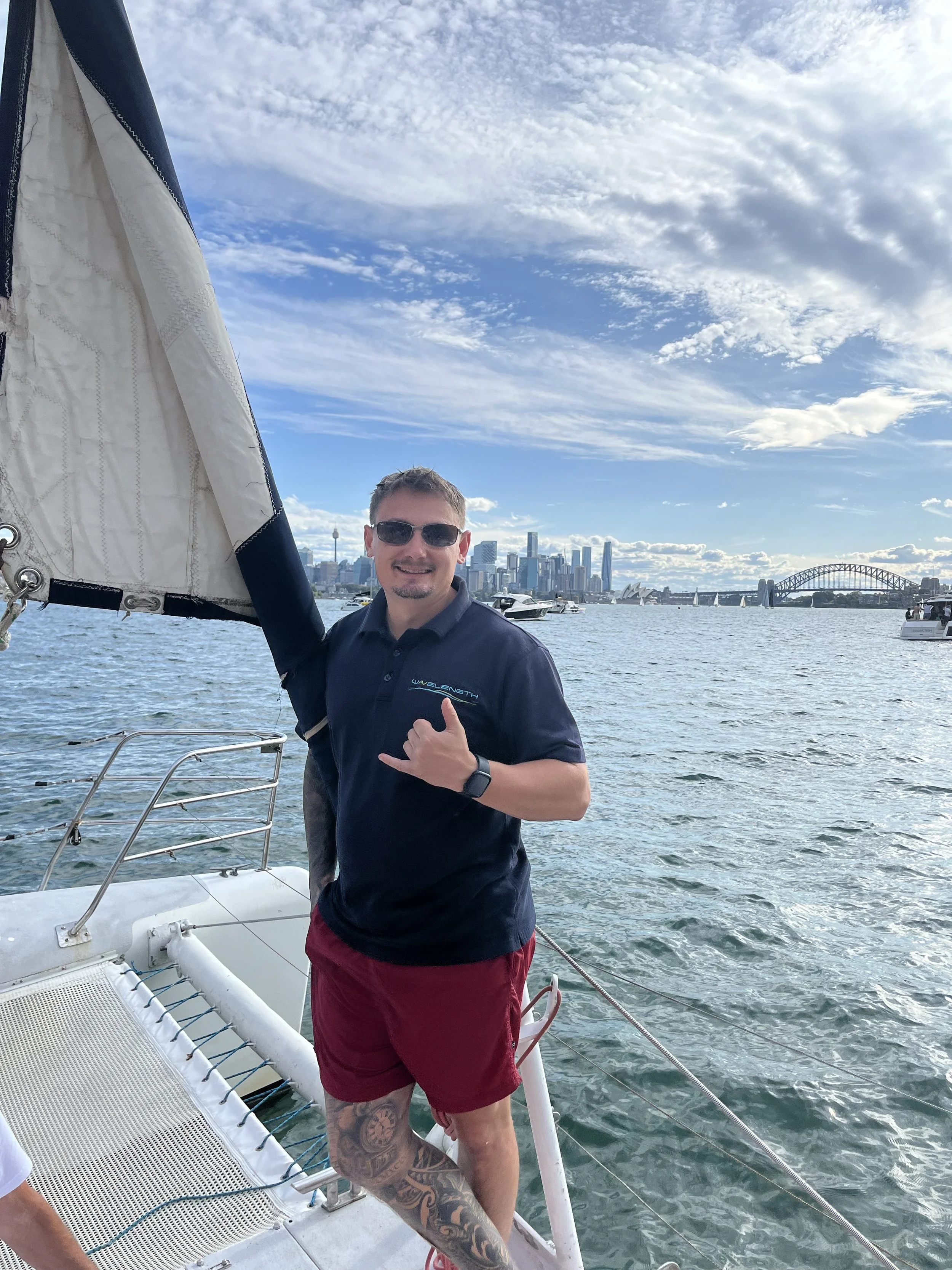 A man standing on a sailboat, wearing sunglasses, a navy polo shirt, and red shorts, with a city skyline and the Sydney Harbour Bridge in the background.