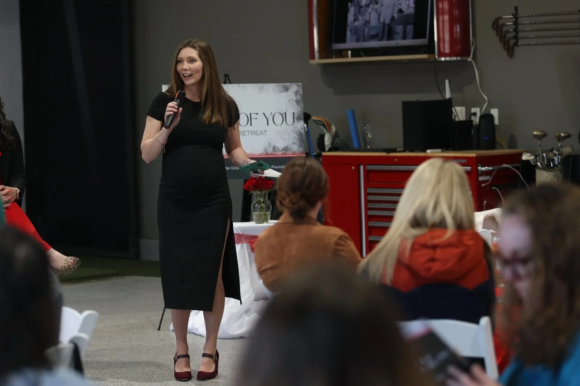 A woman in a black dress with a high slit speaking into a microphone during a presentation or event, with seated audience members around her.