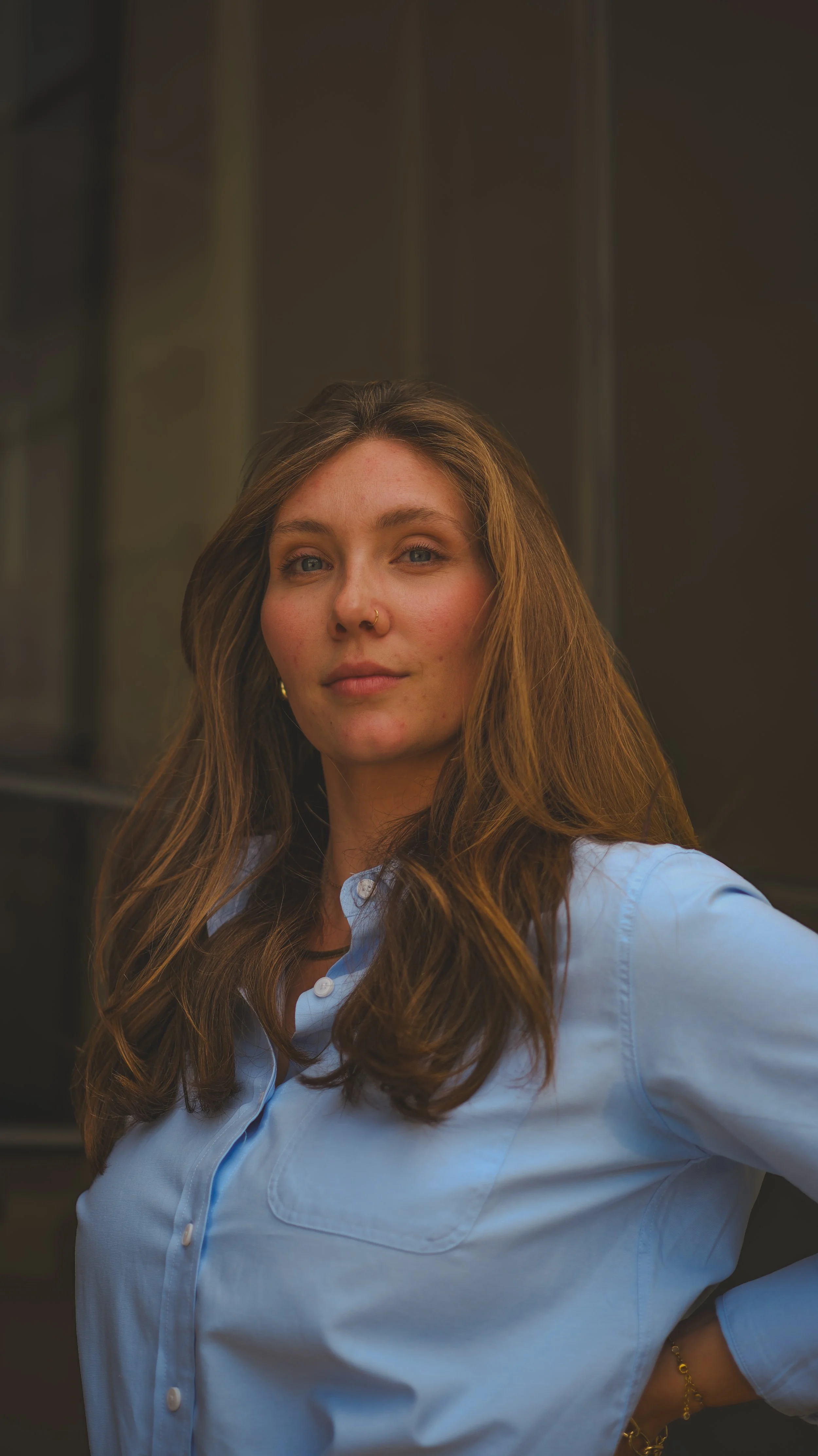 Close-up of a young woman with long wavy brown hair, wearing a light blue button-up shirt, and a gold bracelet, standing outdoors against a dark blurred background.