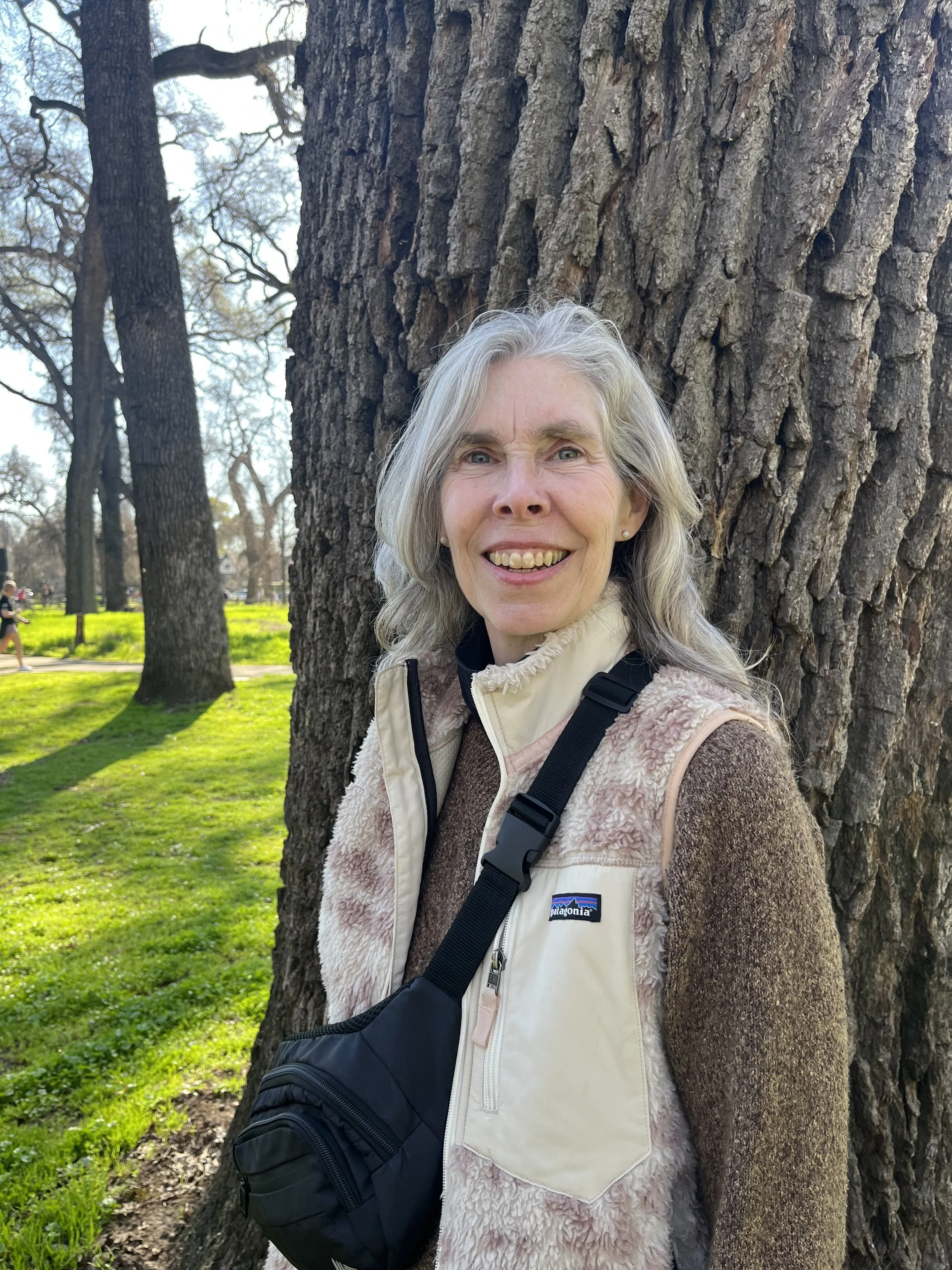 A woman with gray hair smiling while standing next to a large tree trunk in a park on a sunny day.