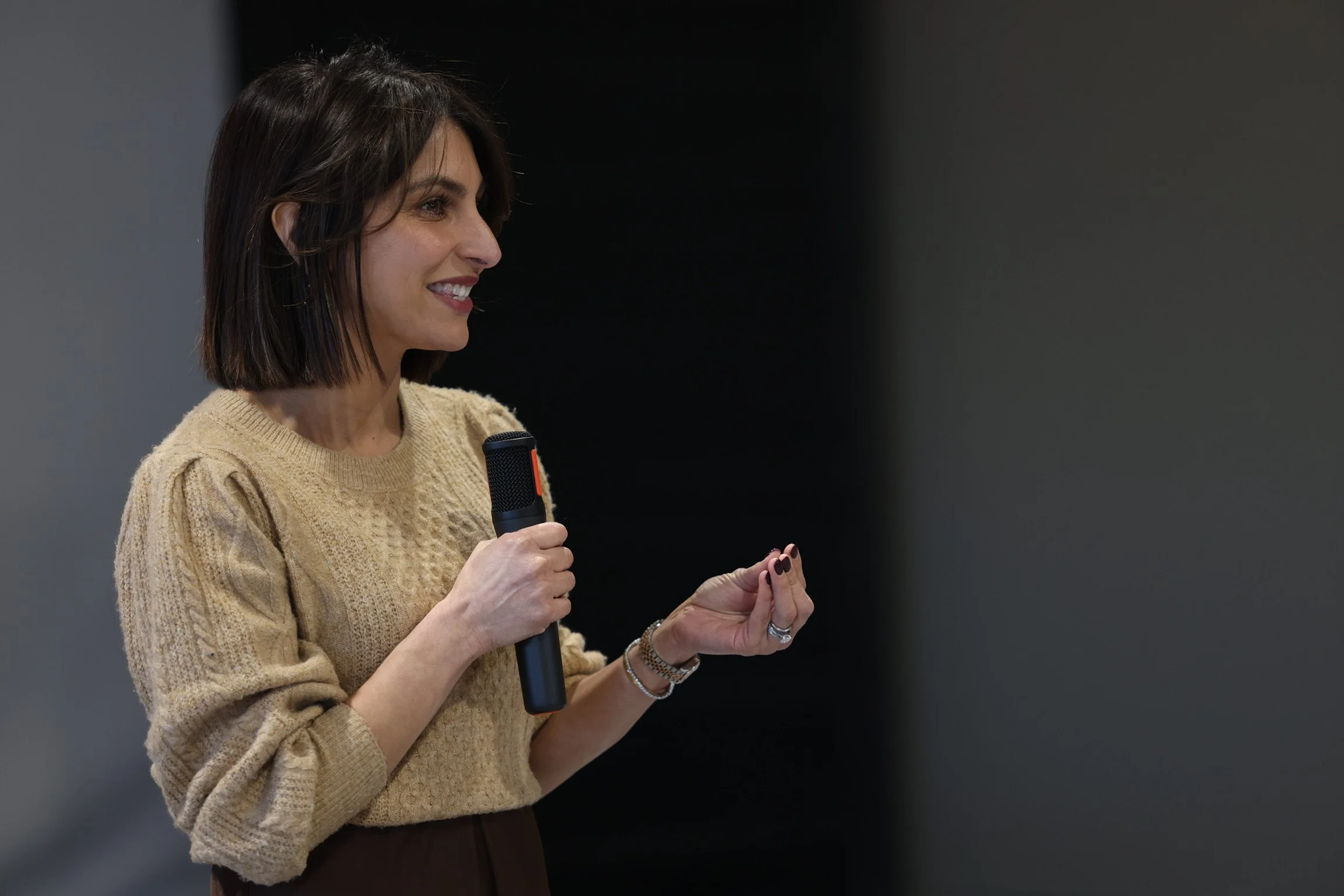 A woman with shoulder-length dark hair and light skin, smiling, holding a microphone in her right hand and gesturing with her left hand, wearing a beige knit sweater.