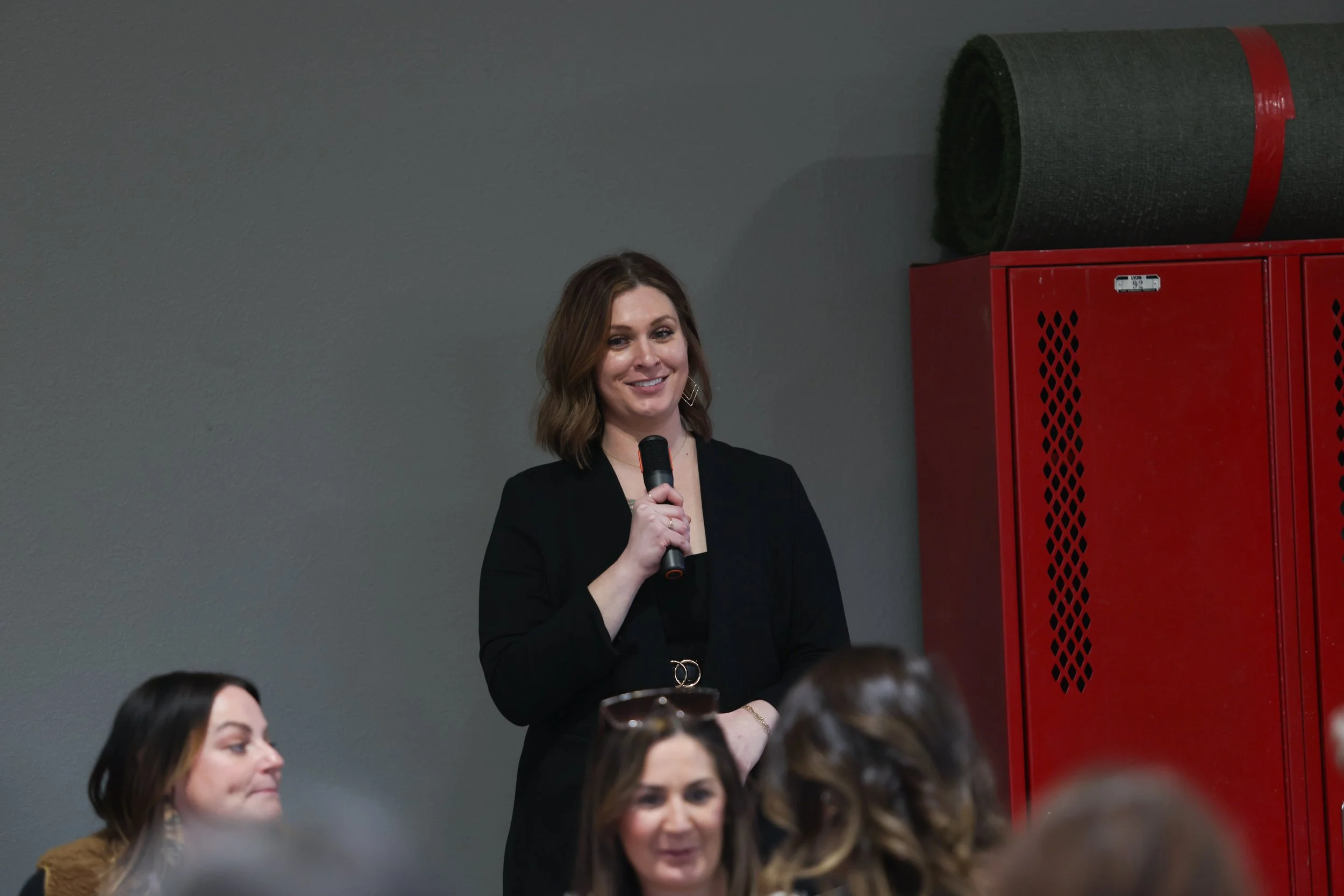 A woman with shoulder-length brown hair, dressed in black, holding a microphone and speaking at an indoor event, with several seated women in front of her and a red locker and large rolled-up mat on the wall behind her.