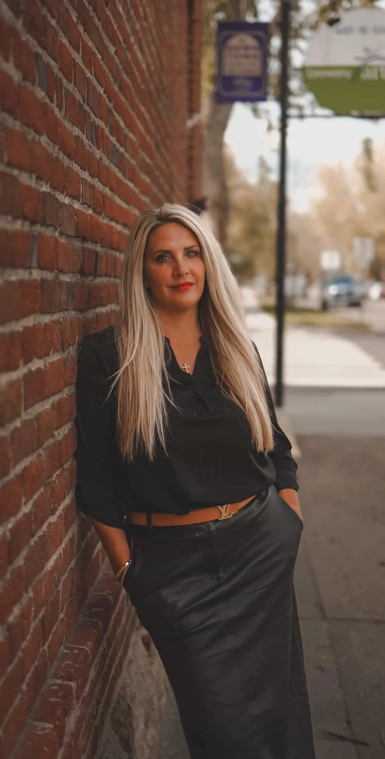 A woman with long blonde hair, red lipstick, wearing a black blouse, leather skirt, and a Louis Vuitton belt, leaning against a brick wall on a city sidewalk.