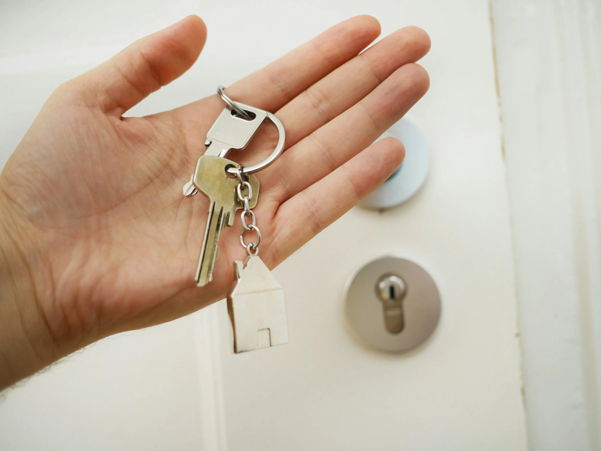 A hand holding a set of house keys with a small house-shaped keychain in front of a white door with a door lock and handle referencing Estate Planning.