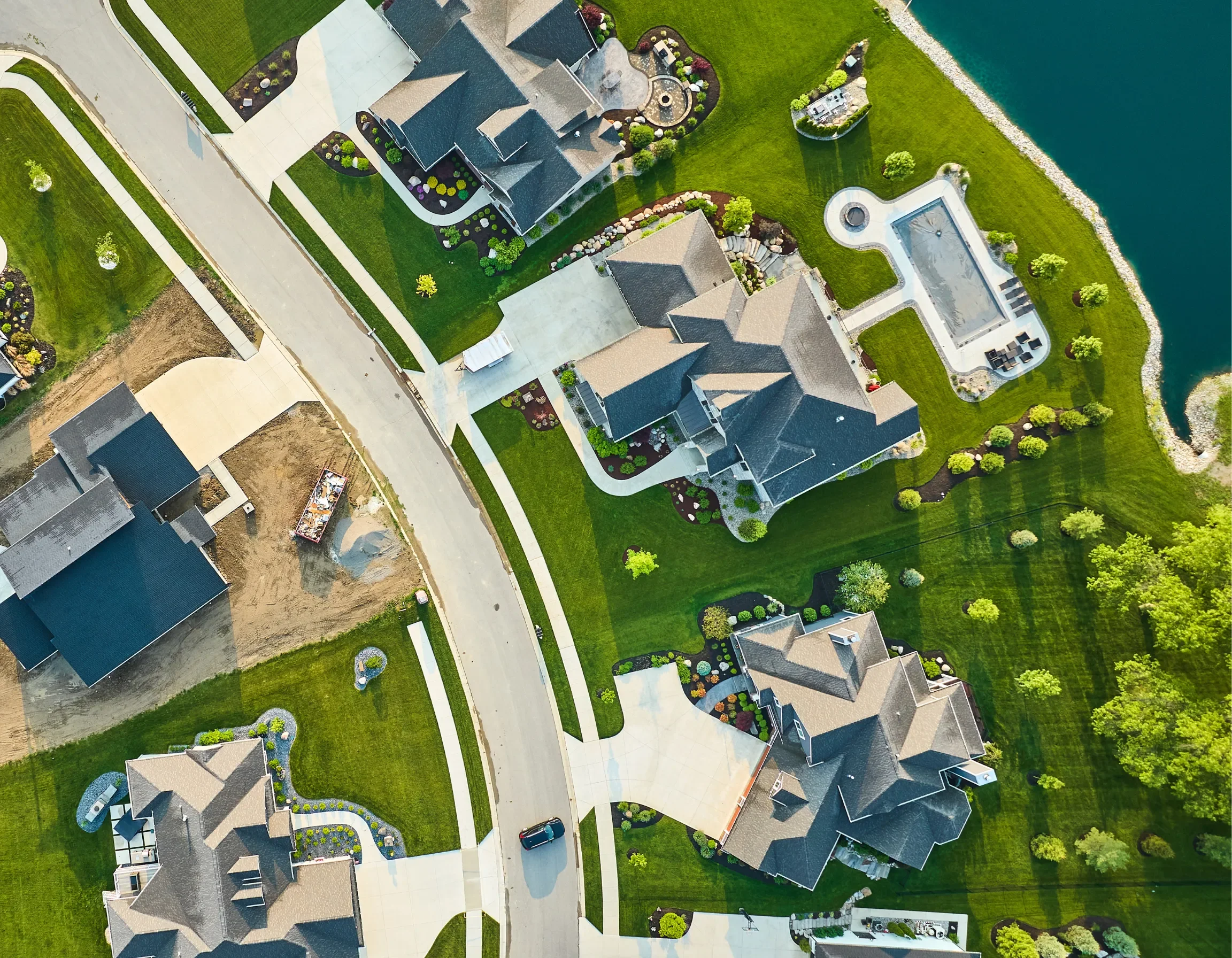 Aerial view of a neighborhood with houses, green lawns, trees, a water body, and a backyard with a pool.