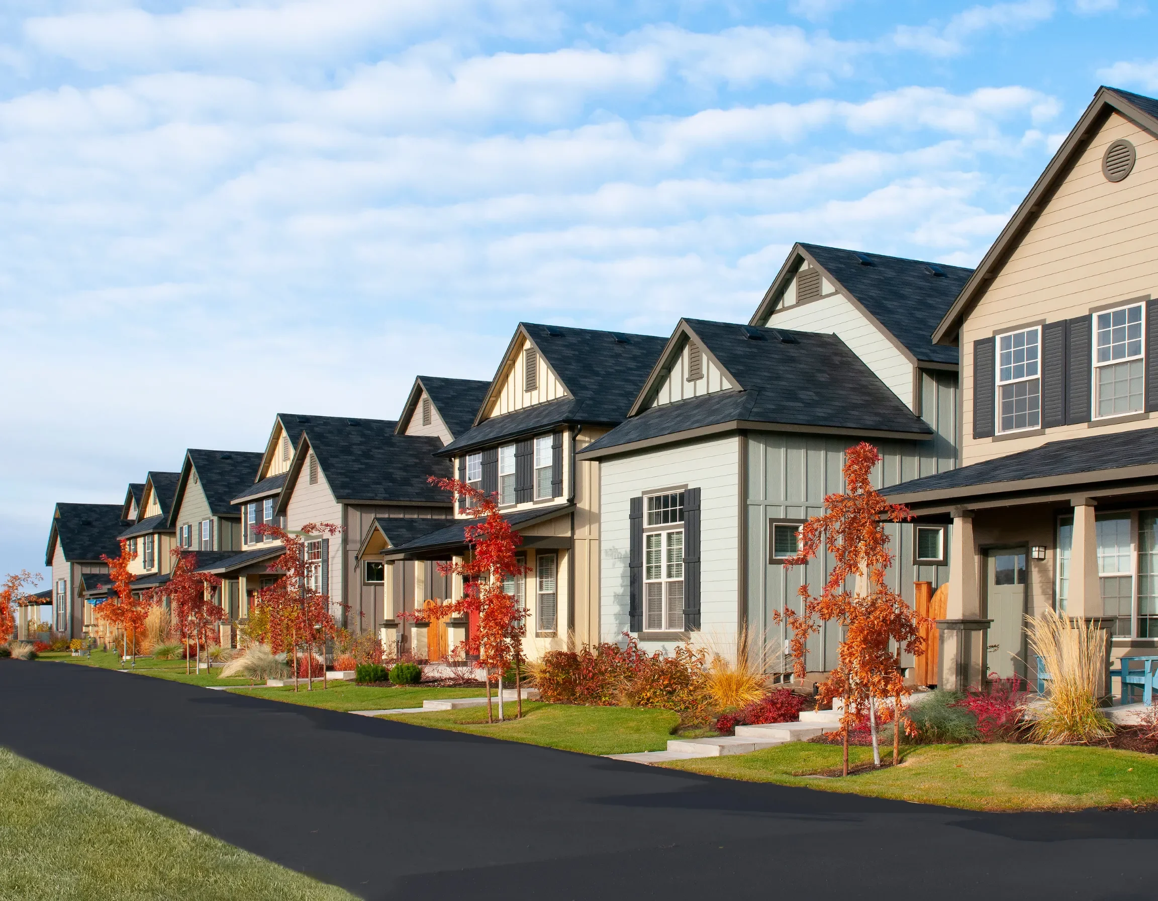 A row of modern residential houses with dark roofs and light-colored siding, lined along a paved street. The front yards feature small trees with red and orange foliage, well-maintained green lawns, and landscaped gardens. The sky above is partly cloudy with blue patches.