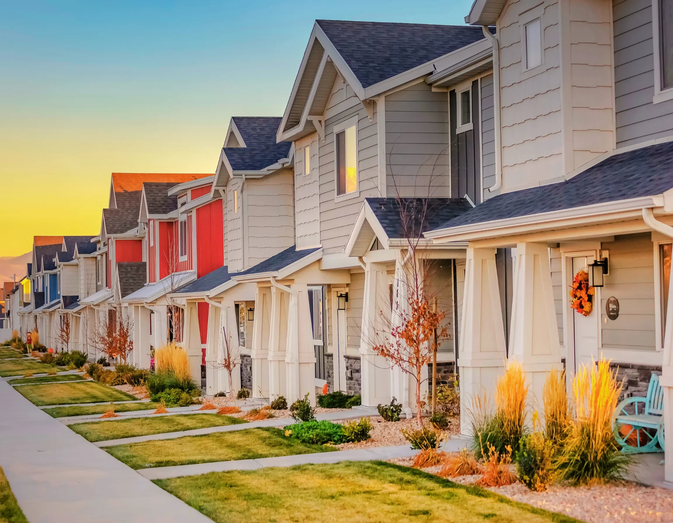 Row of modern townhouses with colorful accents, front yards, and a sidewalk at sunset