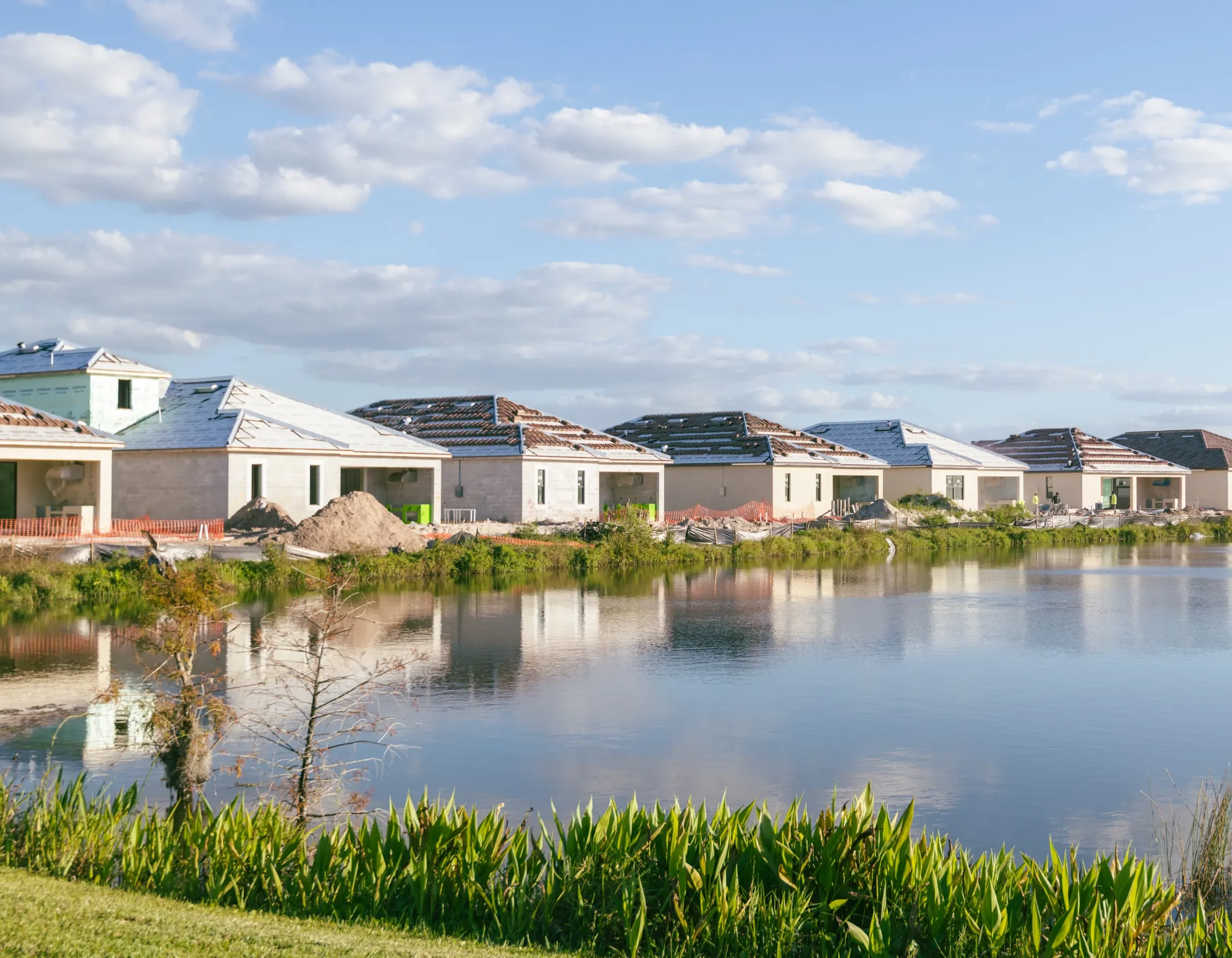 Newly built houses with roofs covered in snow, located next to a water body with green plants in the foreground, under a partly cloudy sky.