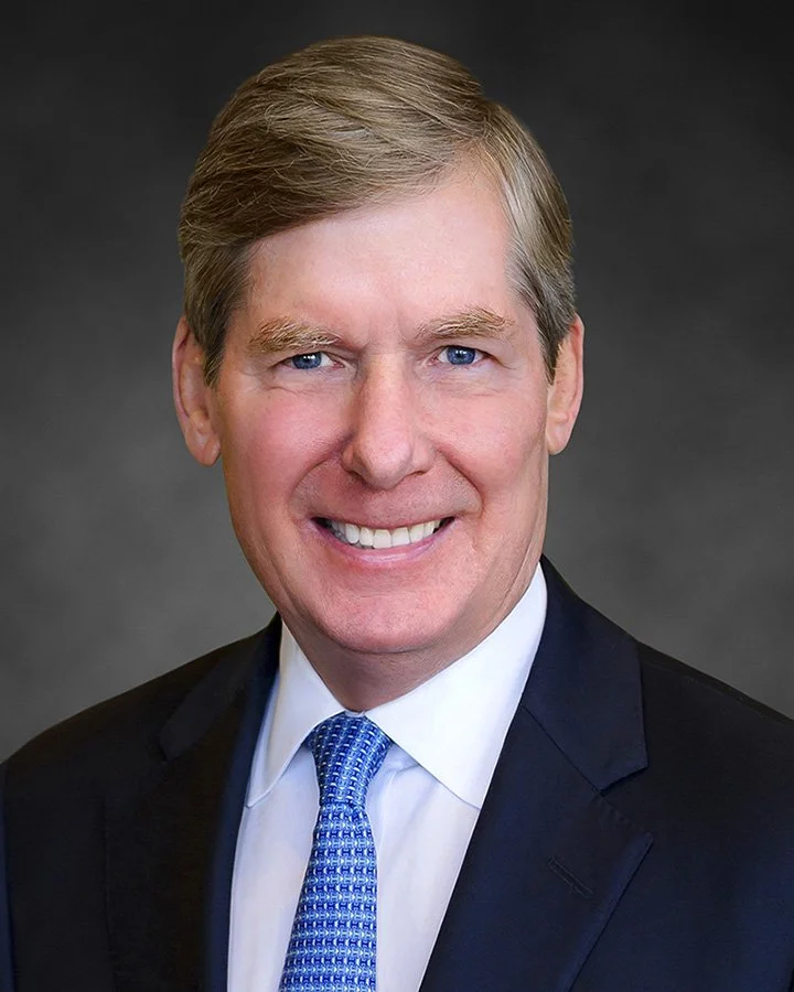 A professional headshot of a man in a suit with a blue tie, smiling against a dark background.