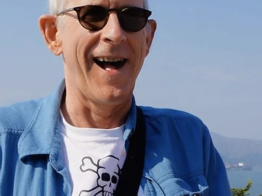 A man wearing sunglasses, a blue shirt, and a white T-shirt with a skull graphic, outdoors with a body of water and mountains in the background, smiling.