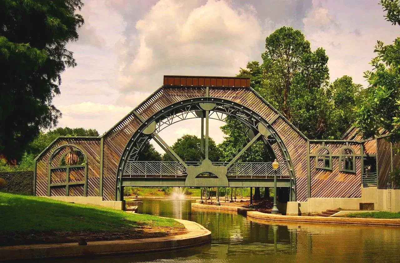 A decorative bridge over a small waterway in a park, surrounded by trees and greenery, with a fountain in the background.