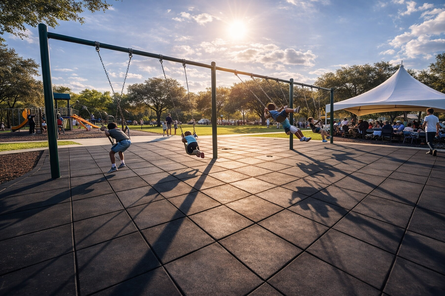 Children playing on swings in a park during sunset, with a group of people sitting under a white tent in the background.