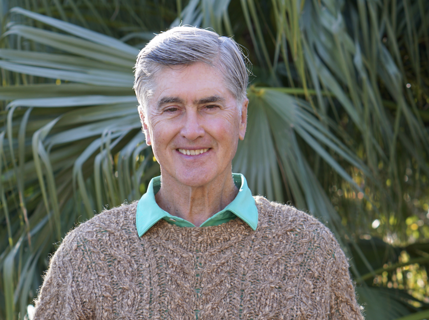 A smiling elderly man with gray hair, wearing a turquoise collared shirt and a brown knitted sweater, standing outdoors in front of large green palm leaves.