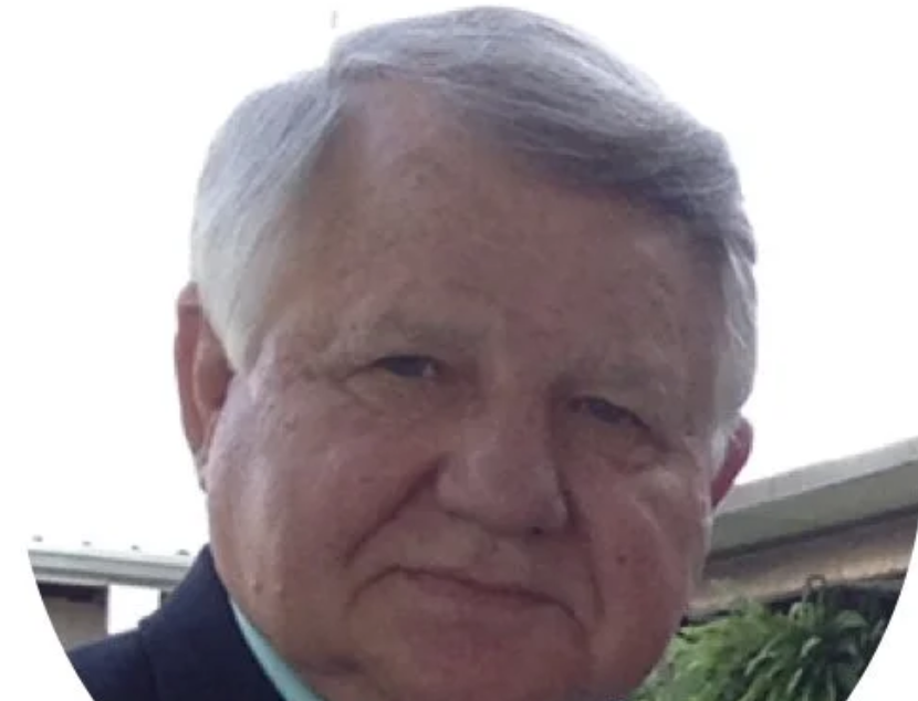 Close-up of an older man with gray hair, wearing a dark suit jacket, outdoors with a background of a building and greenery.