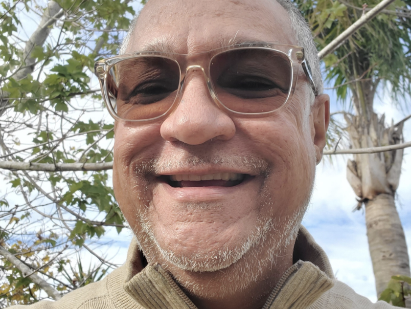 A smiling man wearing sunglasses outdoors with trees and a cloudy sky in the background.