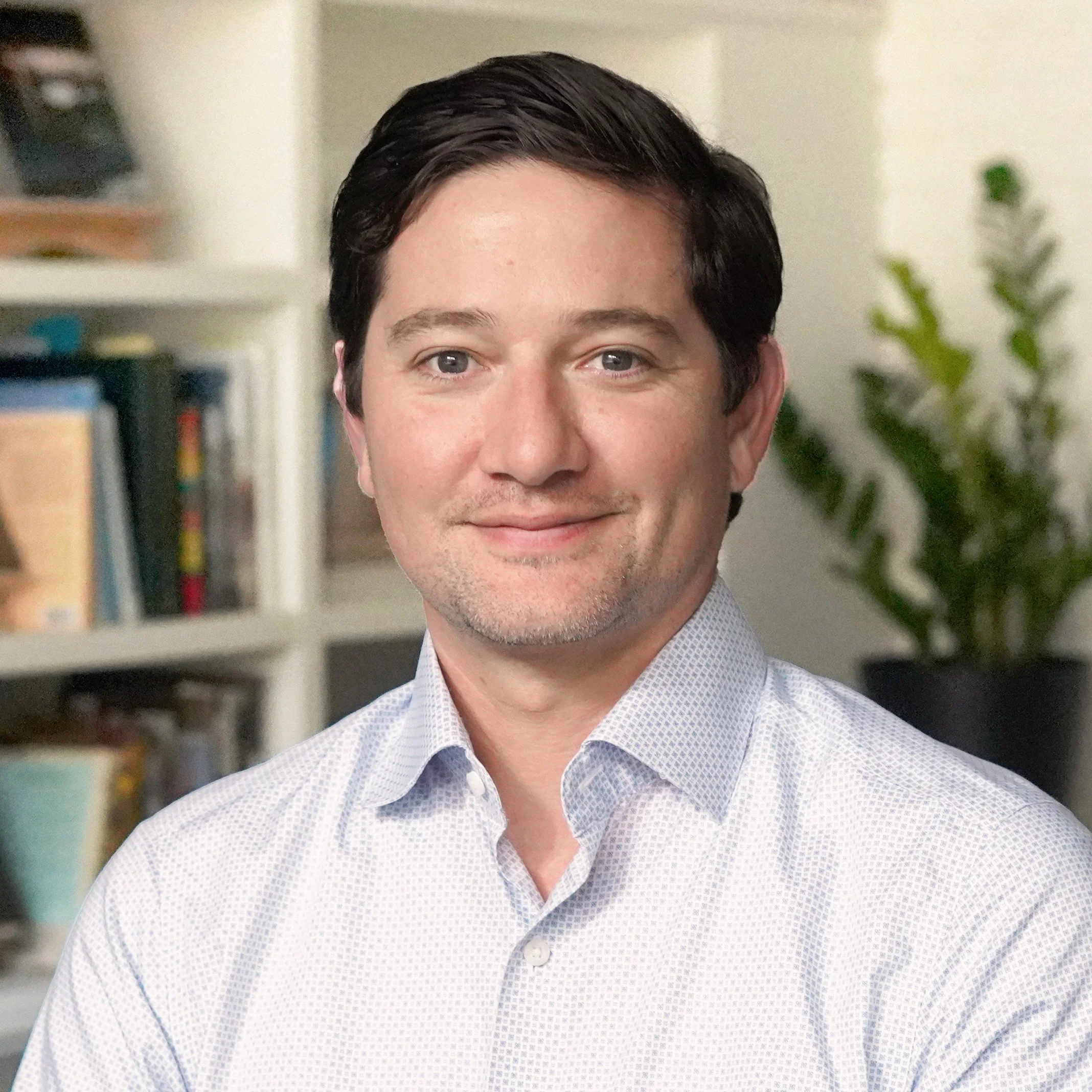A man with dark hair and blue eyes smiling, wearing a light-colored, patterned dress shirt, in a room with bookshelves and a plant in the background.