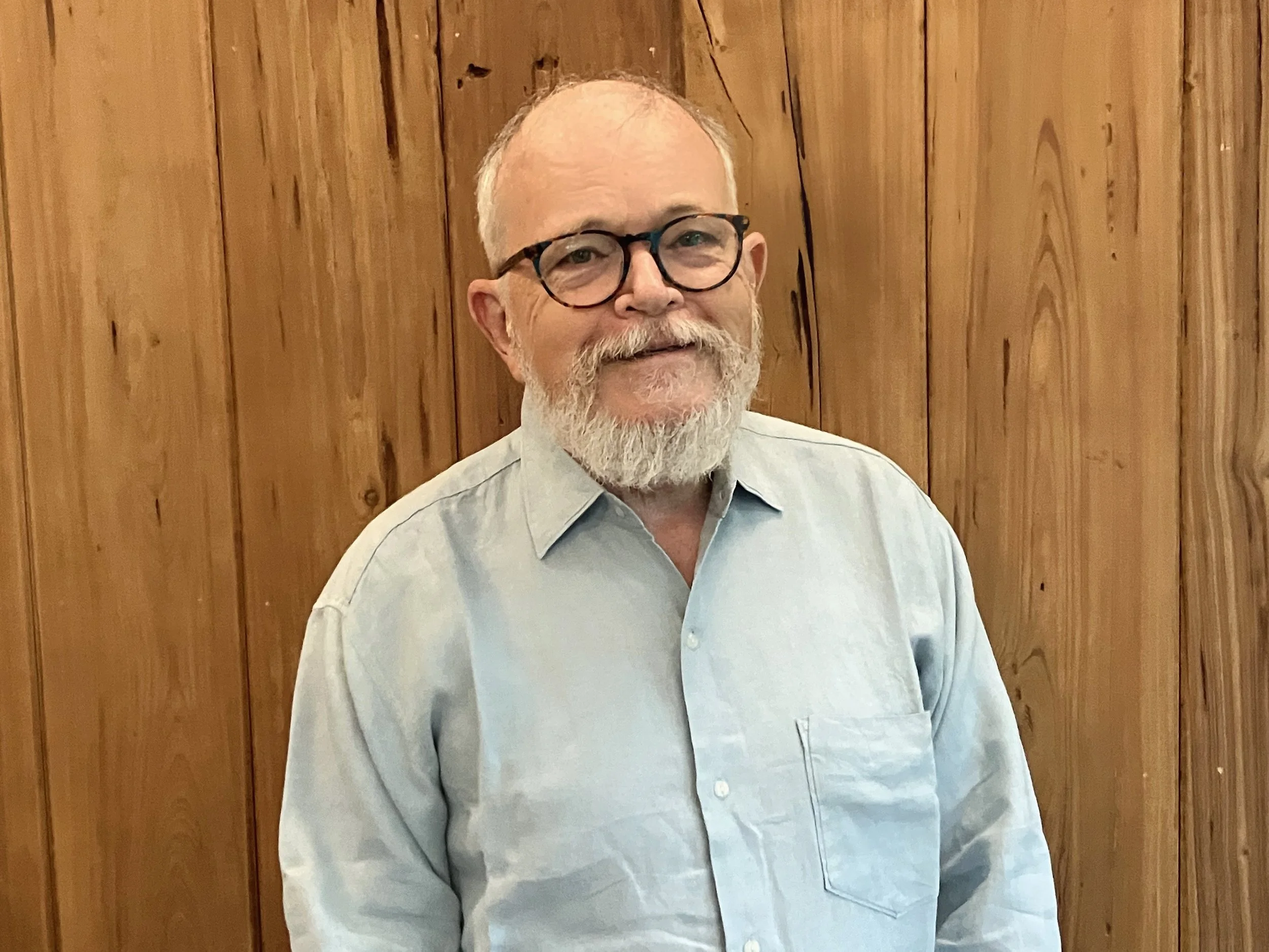A smiling middle-aged man with a beard, glasses, wearing a light-colored shirt, standing in front of a wooden wall.