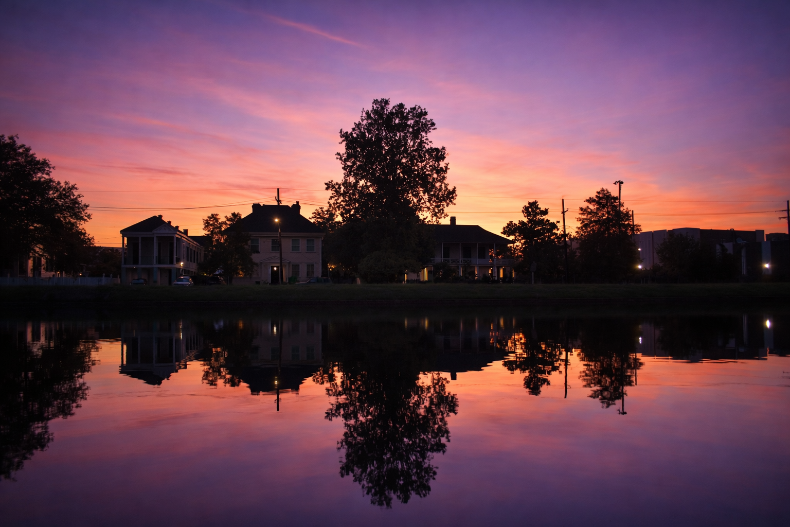 Sunset over a residential neighborhood with houses and trees, reflected in a calm body of water.