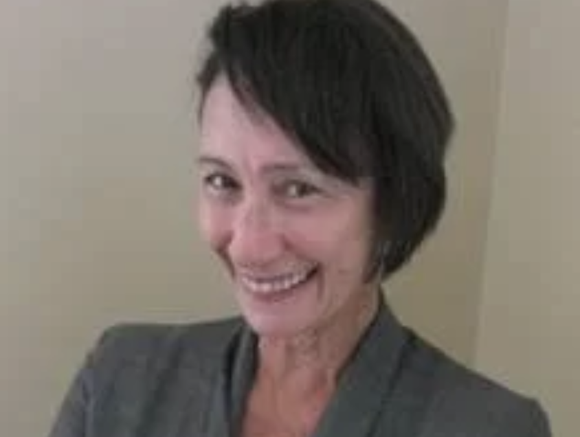 A woman with short dark hair, smiling and wearing a gray blazer, against a plain beige wall.