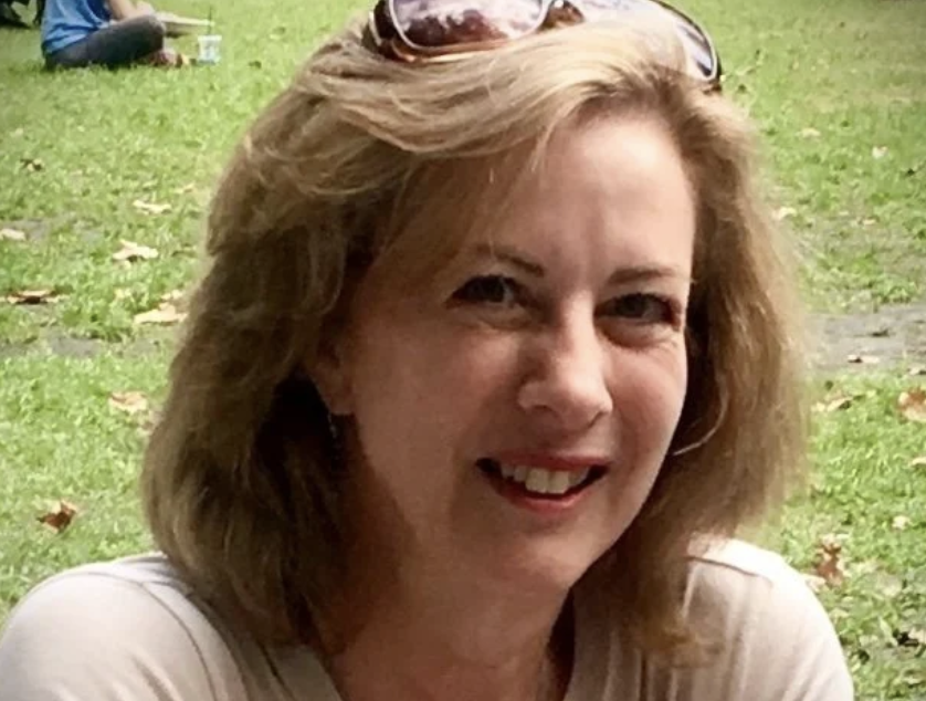 A woman with light brown hair smiling outdoors on a grassy area, with sunglasses on her head and a person sitting in the background.