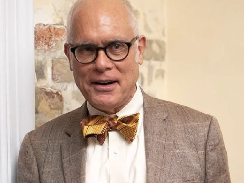 An older man with white hair, glasses, and light skin wearing a brown plaid suit, white shirt, and a yellow plaid bow tie, standing in front of a brick and cream-colored wall.