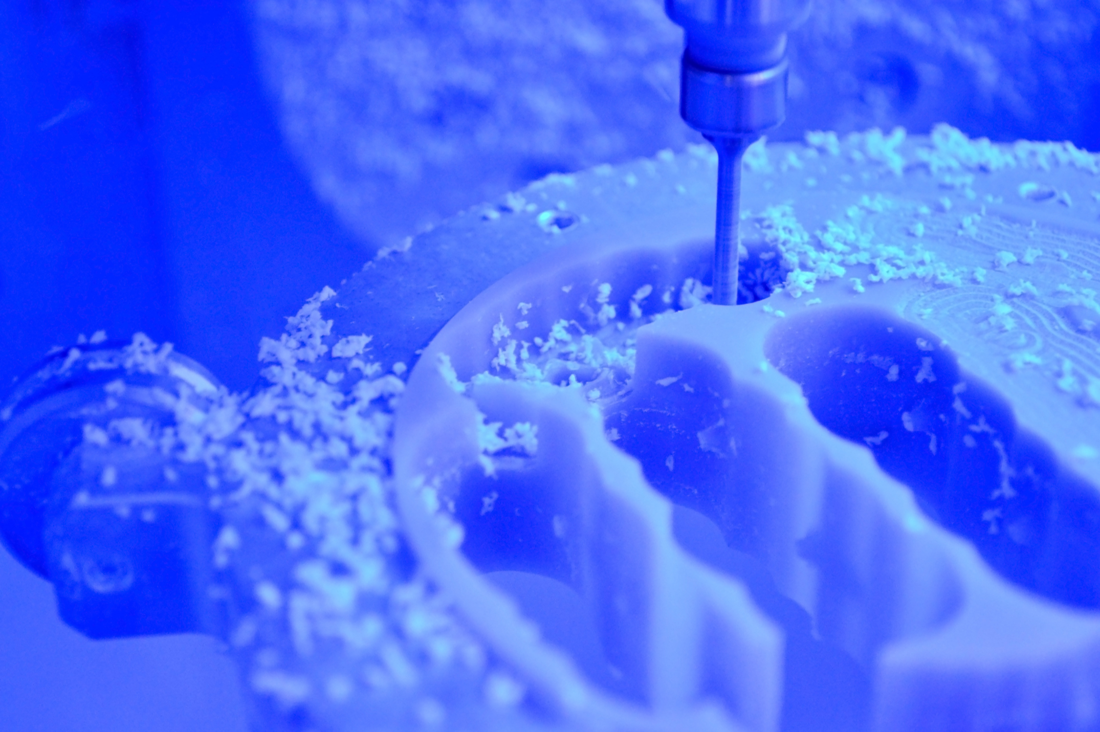 A CNC machine drilling into a white circular object with shavings around it, illuminated by blue light.